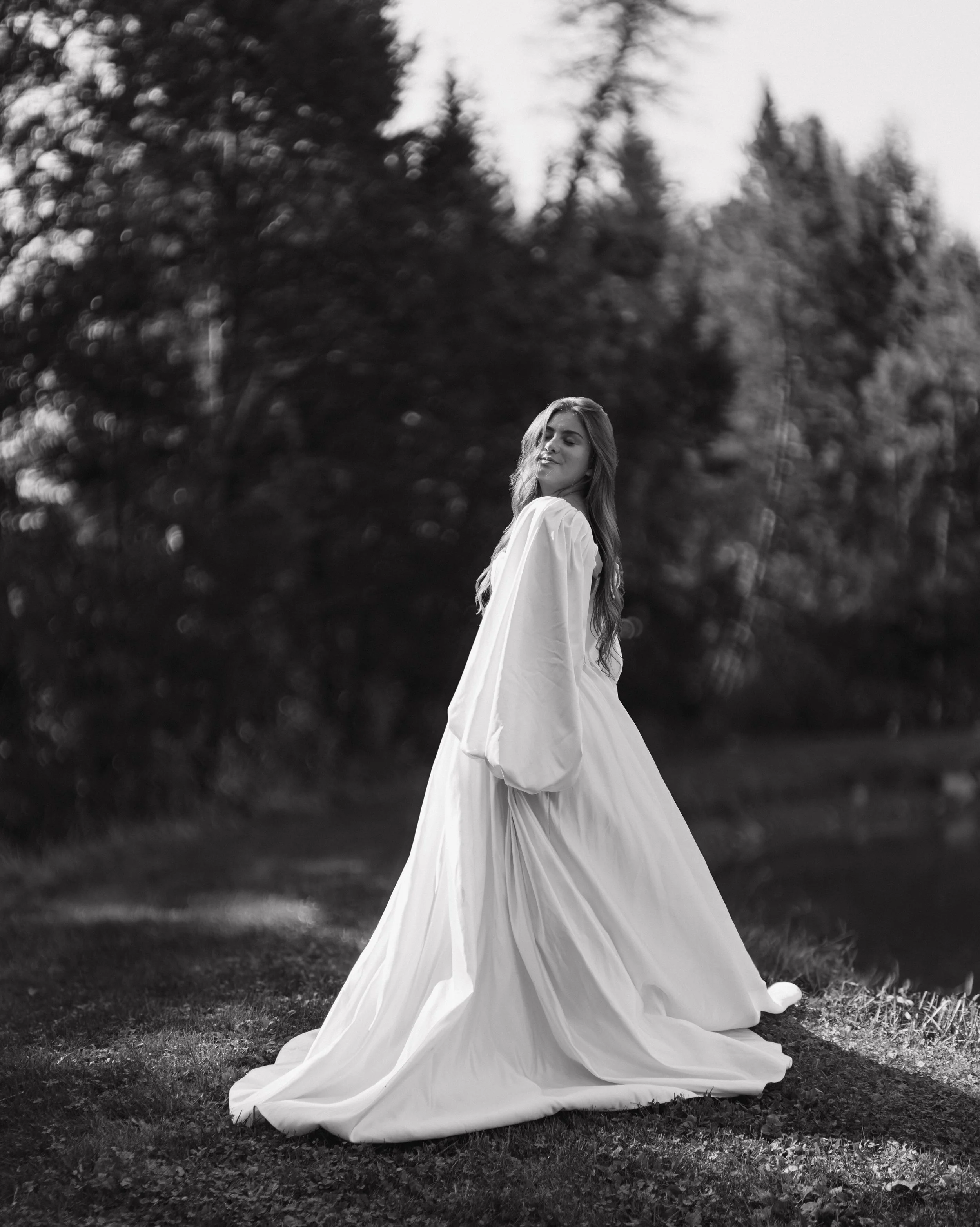 Irasburg Vermont, A woman in a flowing white dress standing outdoors on a grassy area with trees in the background, captured in black and white.