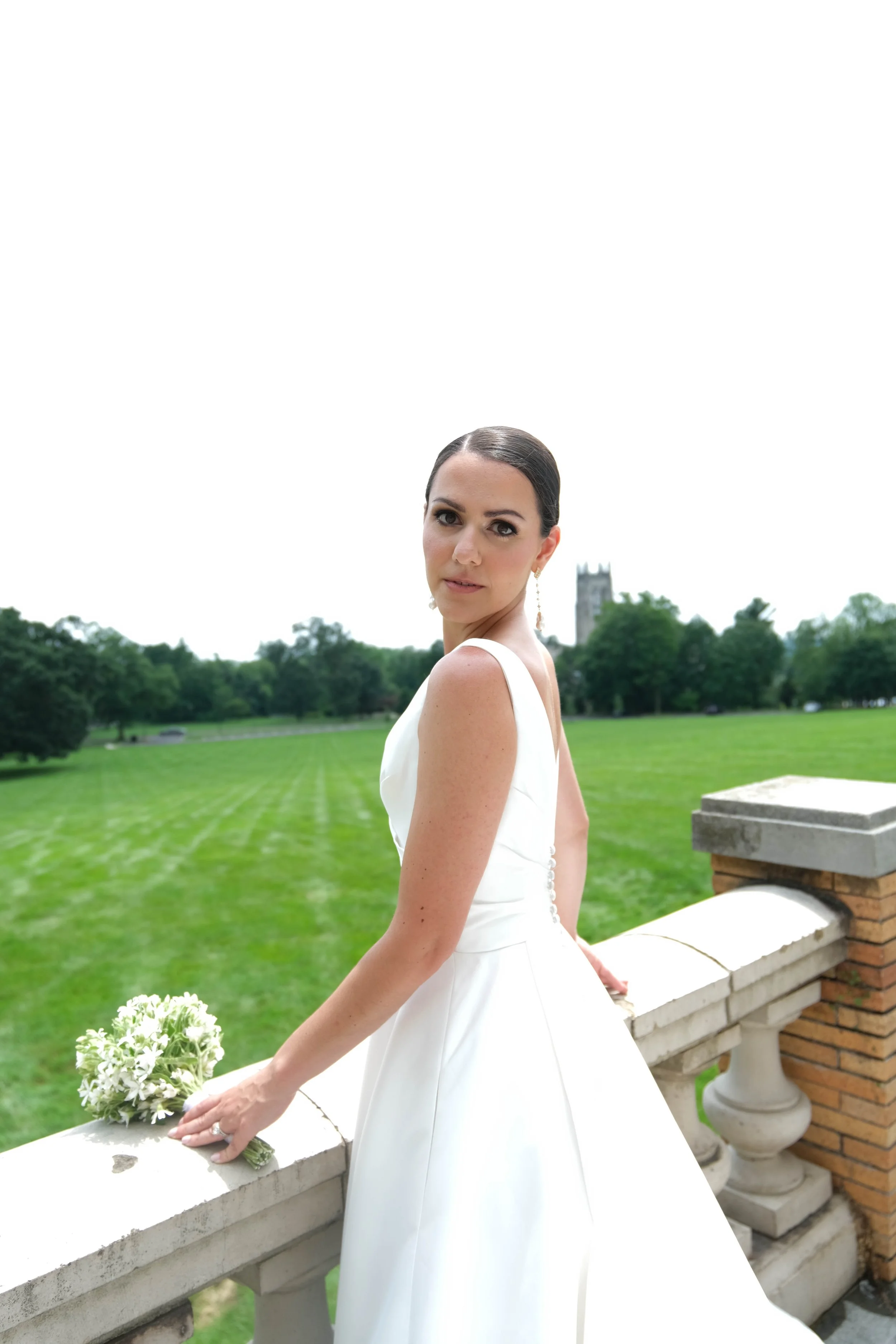 A woman in a white wedding dress standing outdoors on a stone balcony with a green field and trees in the background, holding a bouquet of white flowers.