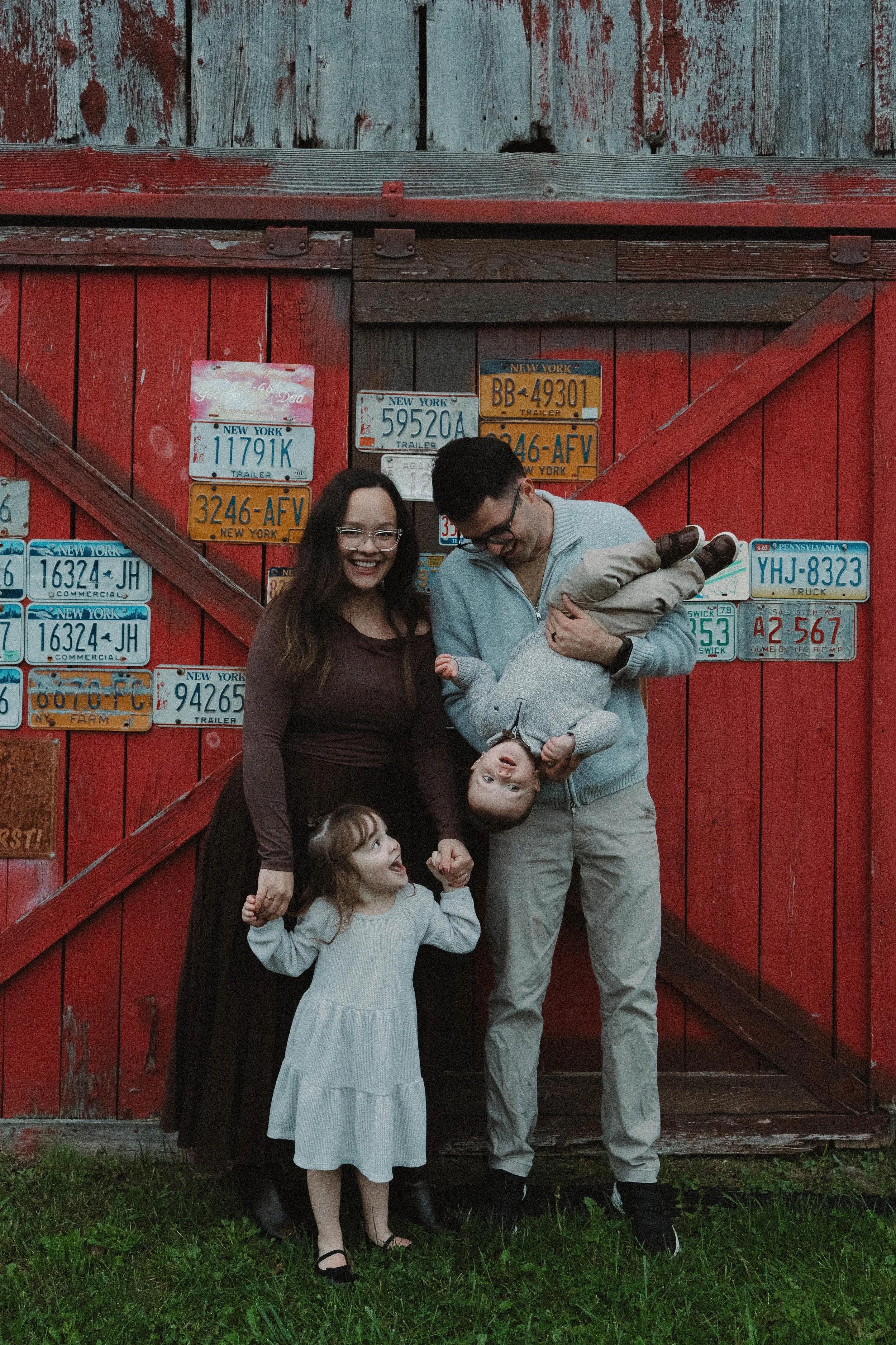 Baldwinsville, NY Photographer Family of four standing in front of a red wooden barn wall decorated with vintage license plates. The mother, father, and two young children are smiling and laughing, with one child being held upside down by the father.