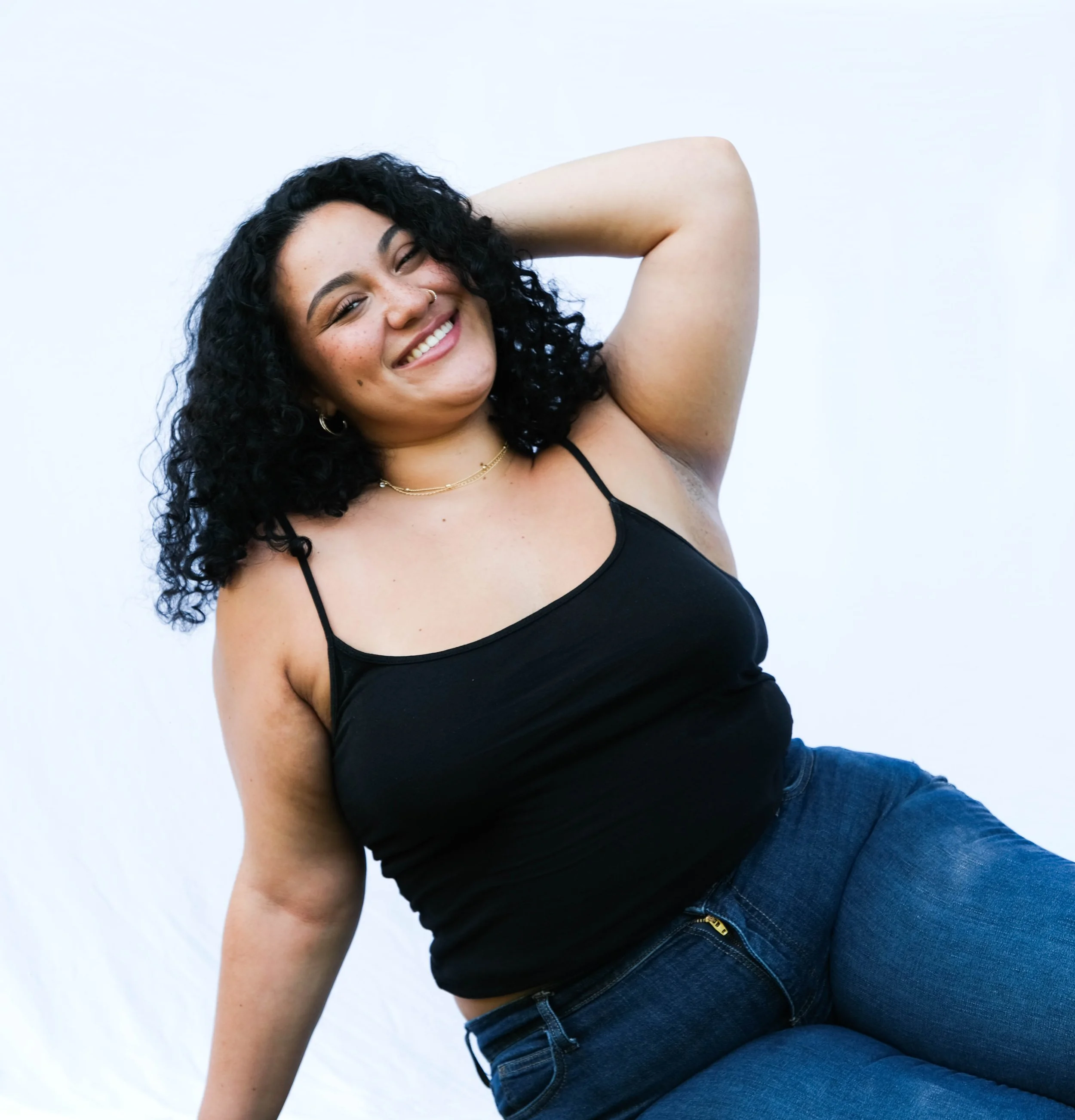 A young woman with curly black hair, wearing a black tank top and blue jeans, smiling and posing with one arm behind her head against a white background.