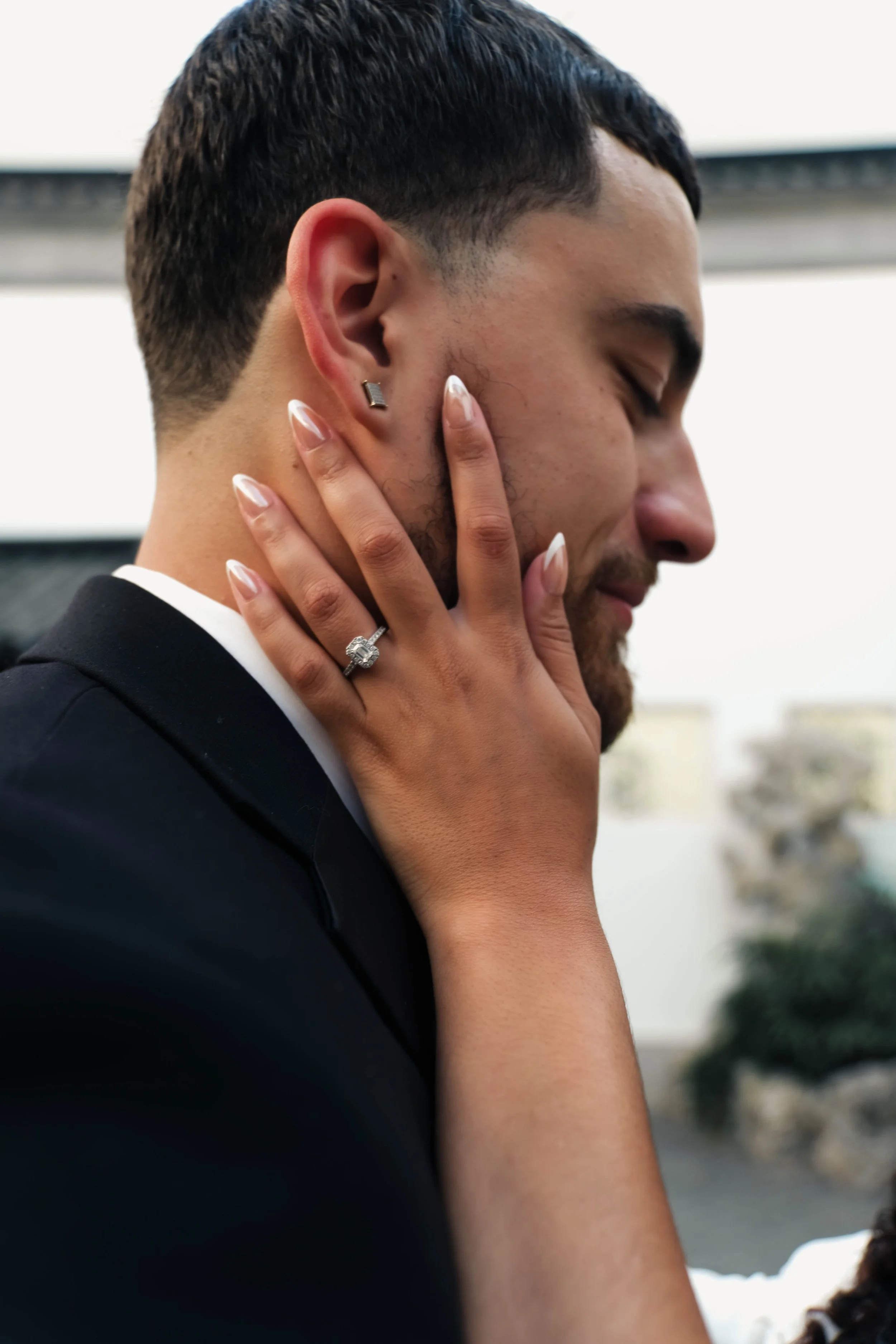 The Met Engagement Shoot  A man in a suit with a beard and earrings is being kissed on the cheek by a woman with manicured nails and a ring, during daytime with a blurry outdoor background.