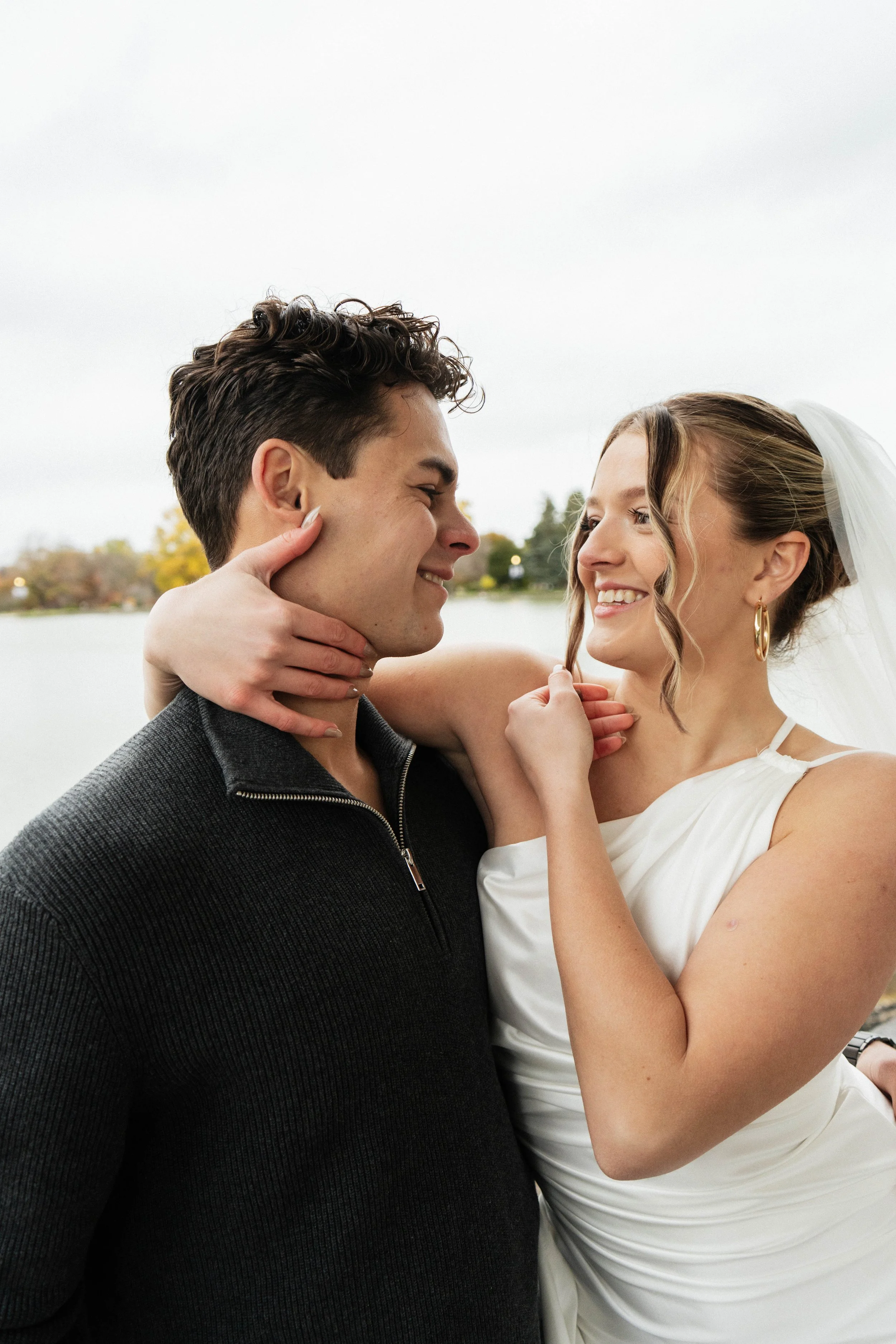 A couple is smiling and lovingly looking at each other outdoors near a body of water, with trees and an overcast sky in the background.