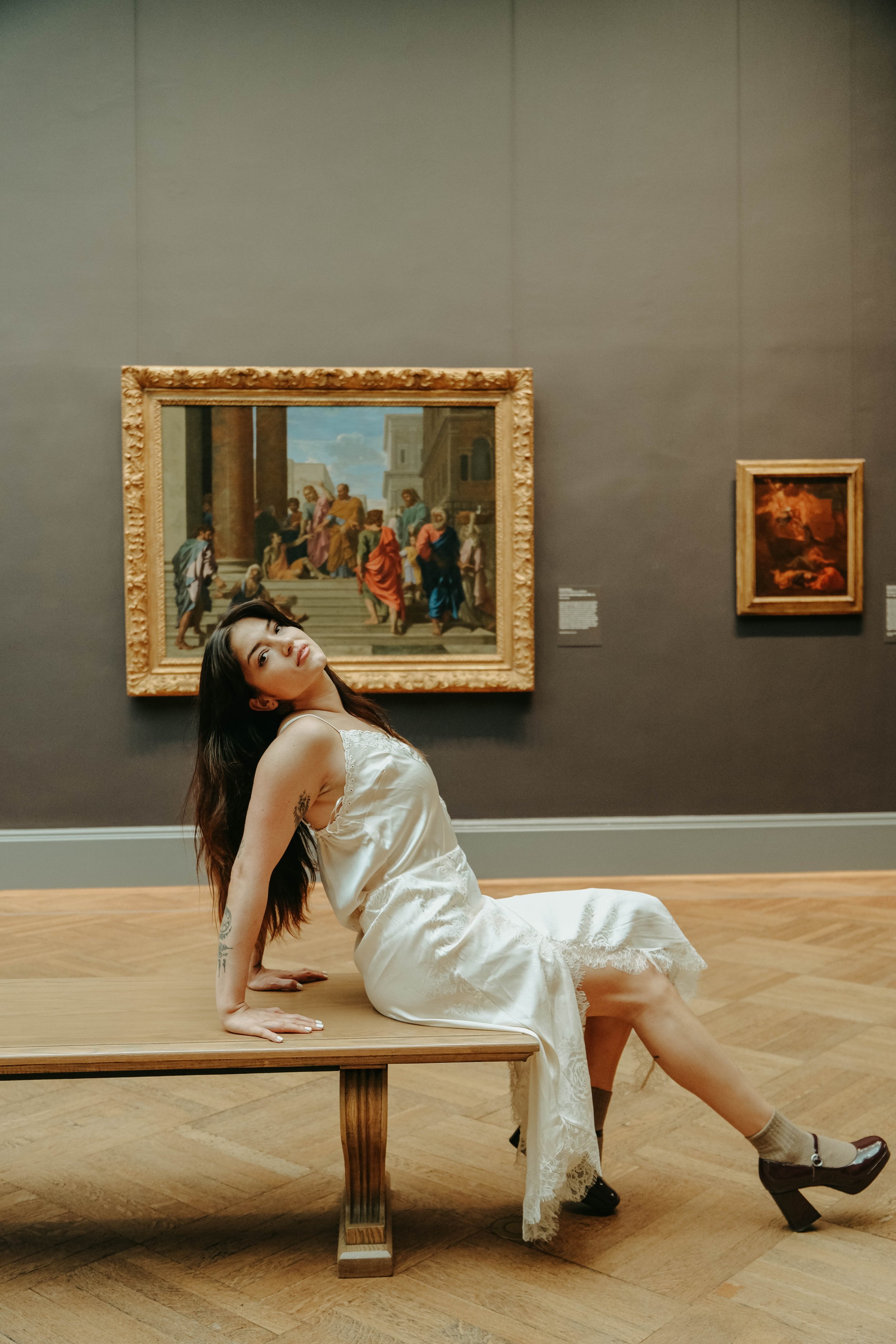 The Mey Museum, A woman in a white dress sitting on a wooden bench in an art gallery with paintings on the wall behind her.