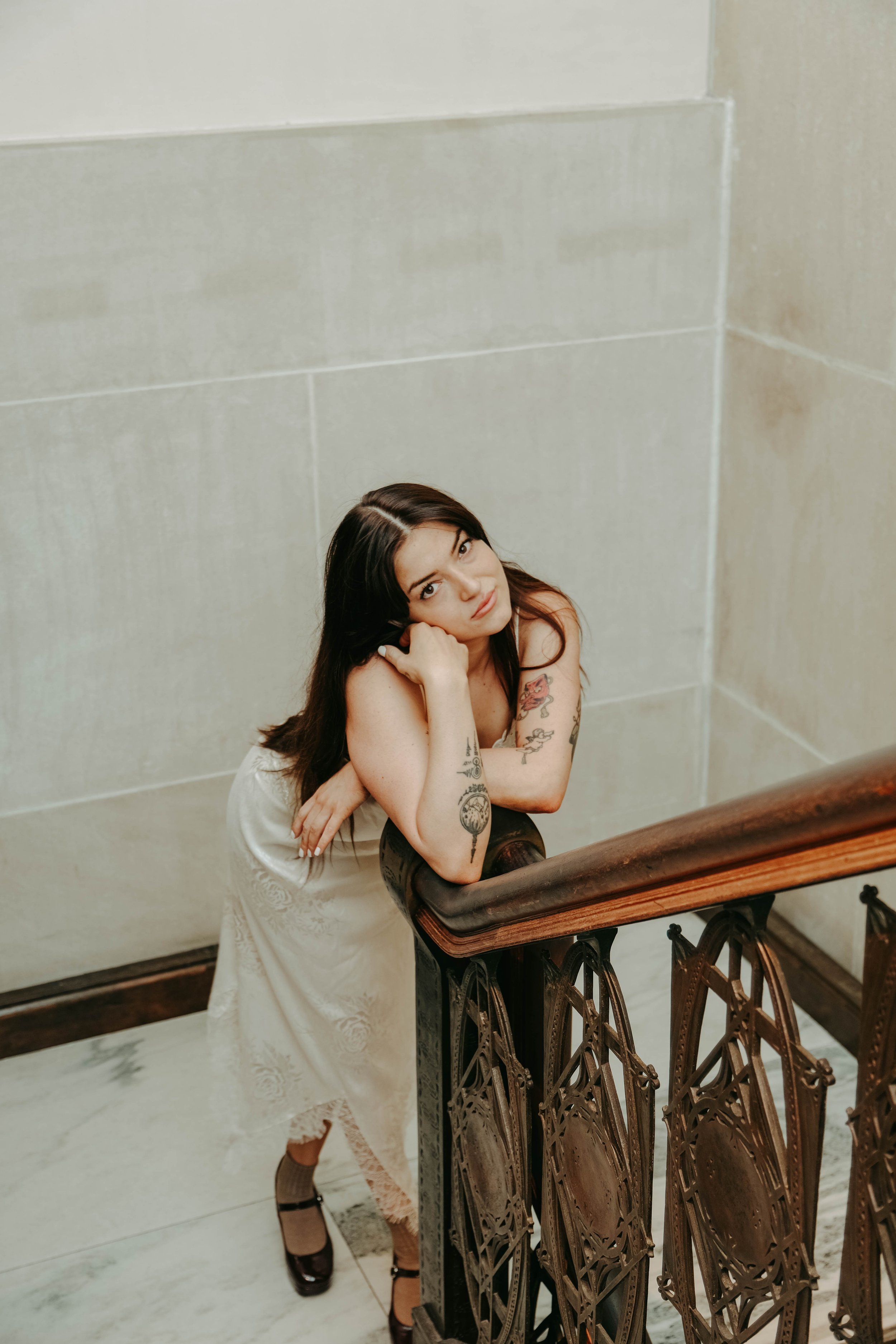 The Met Museum, Young woman with tattoos leaning on a staircase banister in a cream dress, looking at the camera.