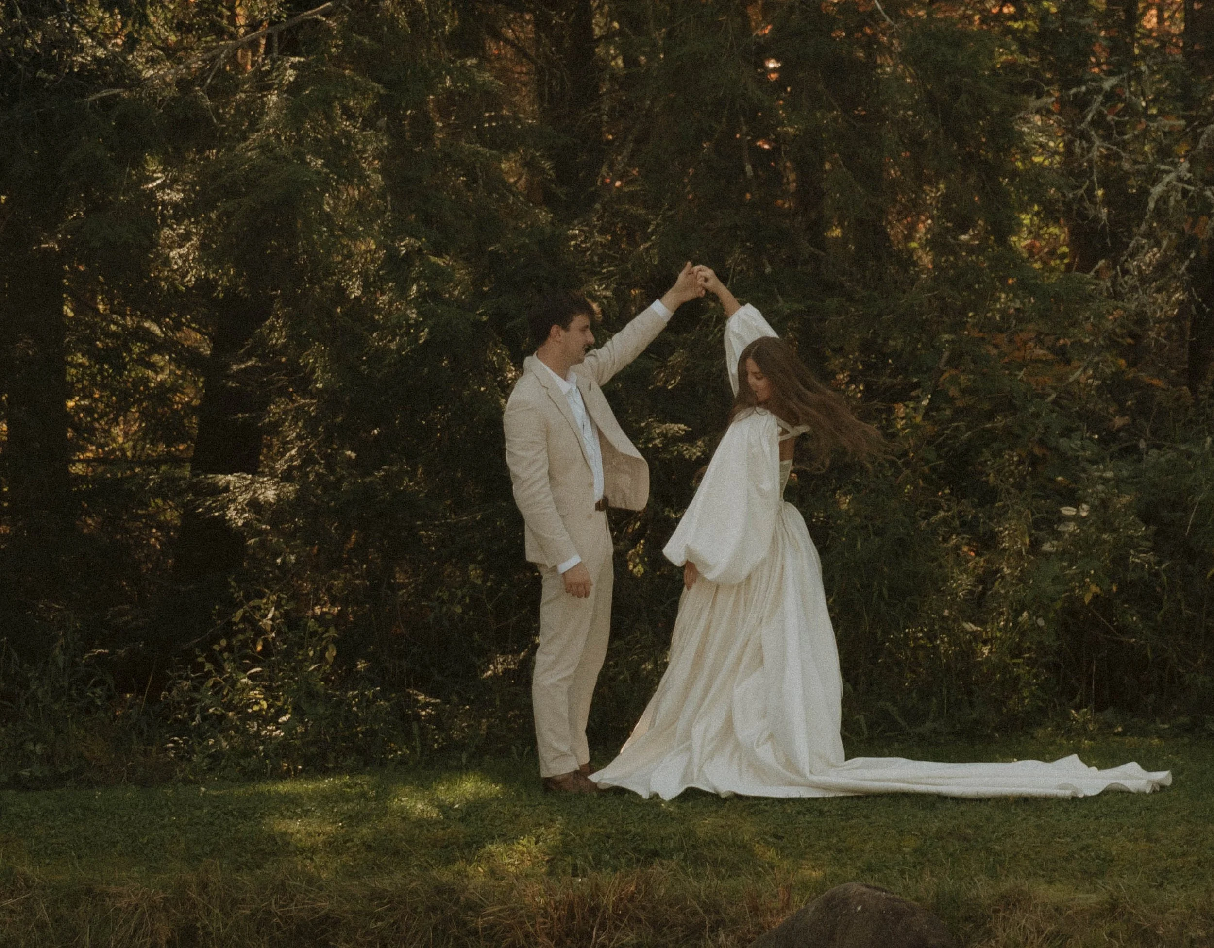 A couple in wedding attire dancing outdoors in a wooded area with sunlight filtering through trees.
