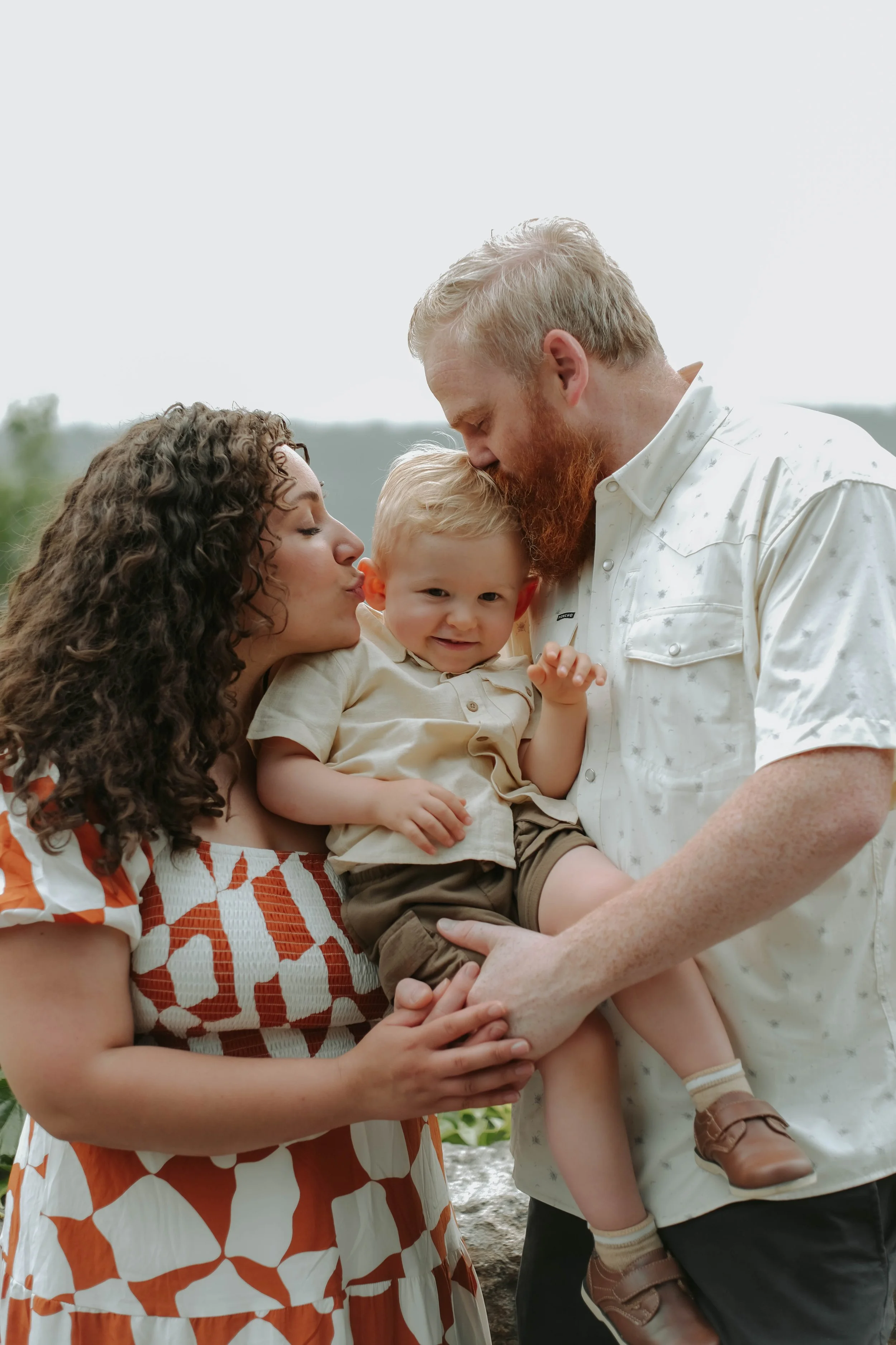 Fort Tryon Park, NYC  A family of three with a mother, father, and young boy outdoors, kissing and holding each other.