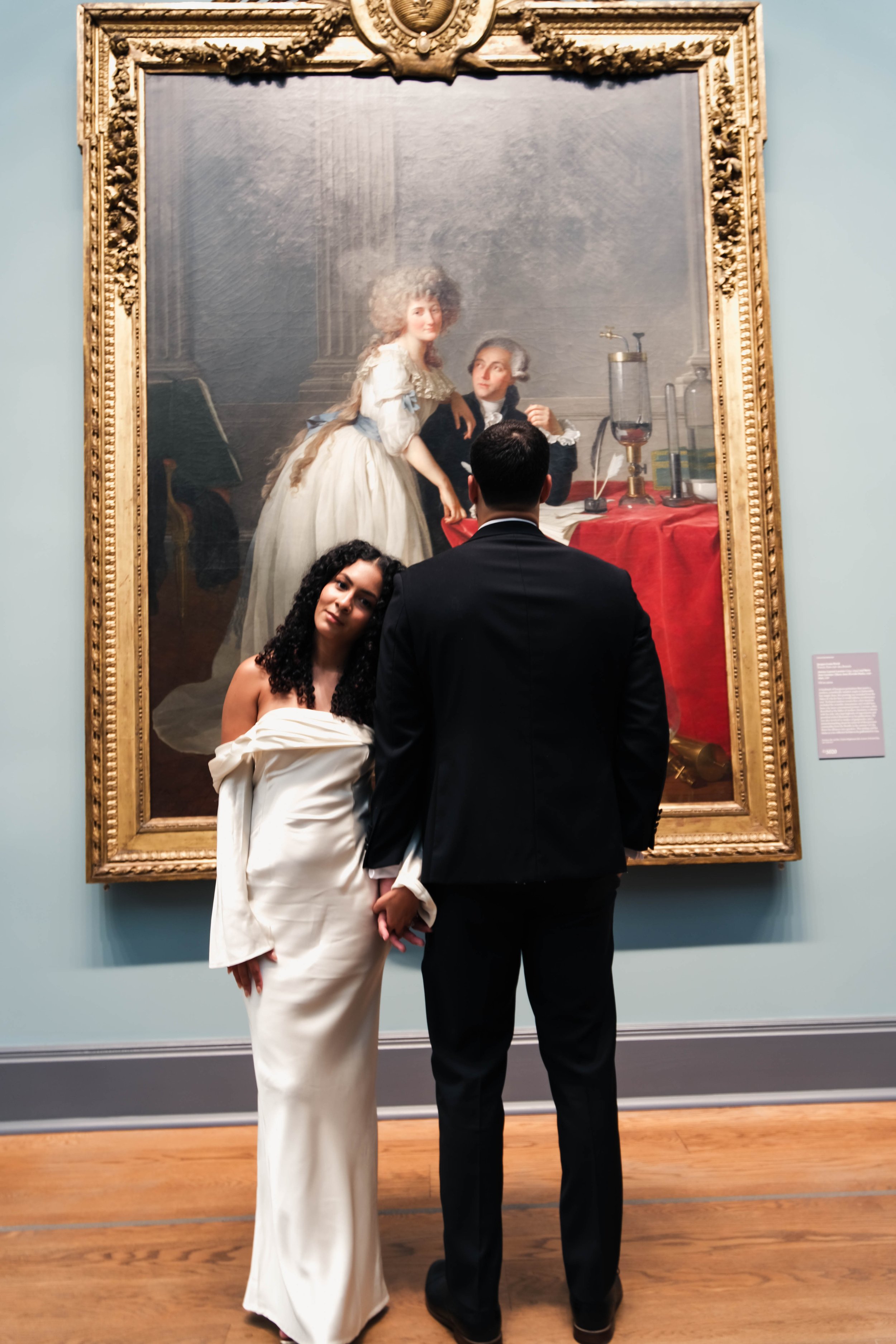 A man in a black suit and woman in a white dress hold hands while standing in front of a large framed classical painting in an art gallery. The painting depicts two historical figures in a scientific context, one seated at a table with scientific equipment and the other standing beside them.