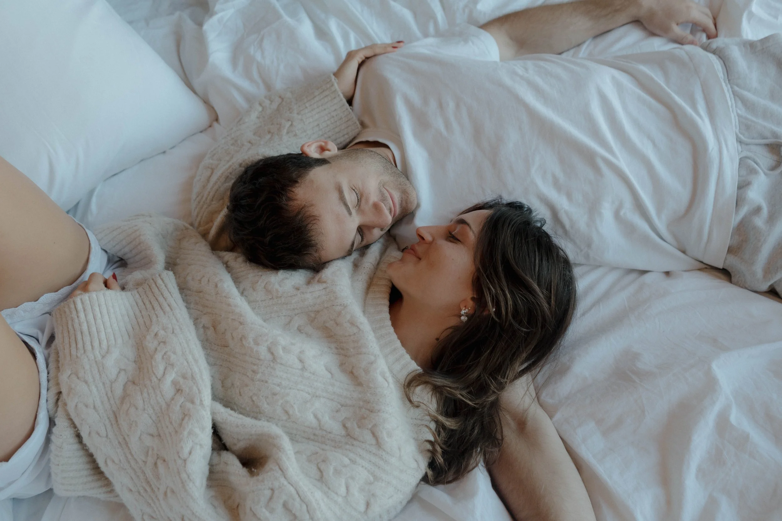 A couple lying in bed, facing each other and smiling softly, sharing an intimate moment.