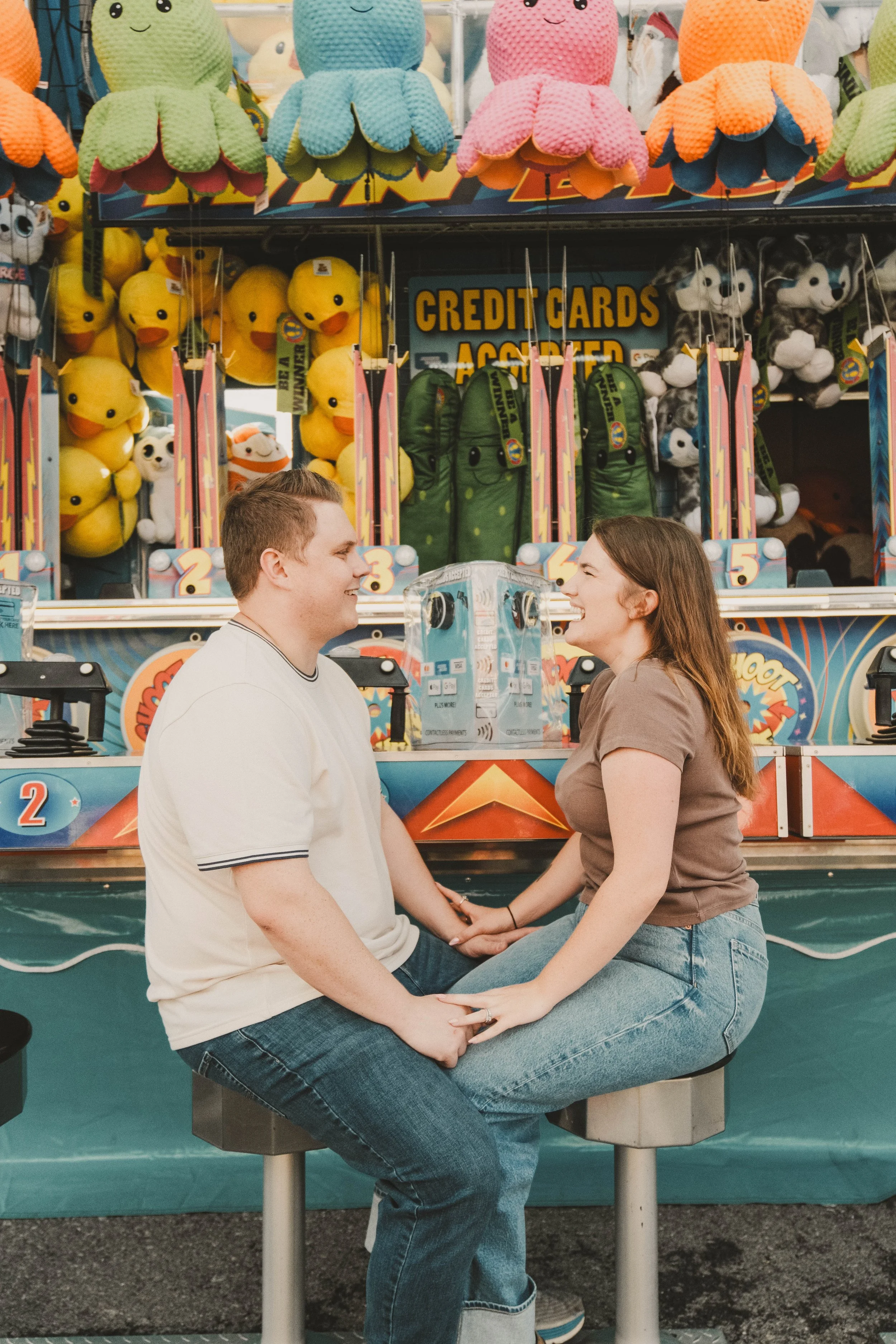 NYS Fair Minis, Syracuse, NY, A young couple sitting on a carousel at a carnival, facing each other and smiling, with arcade game and plush toys in the background.