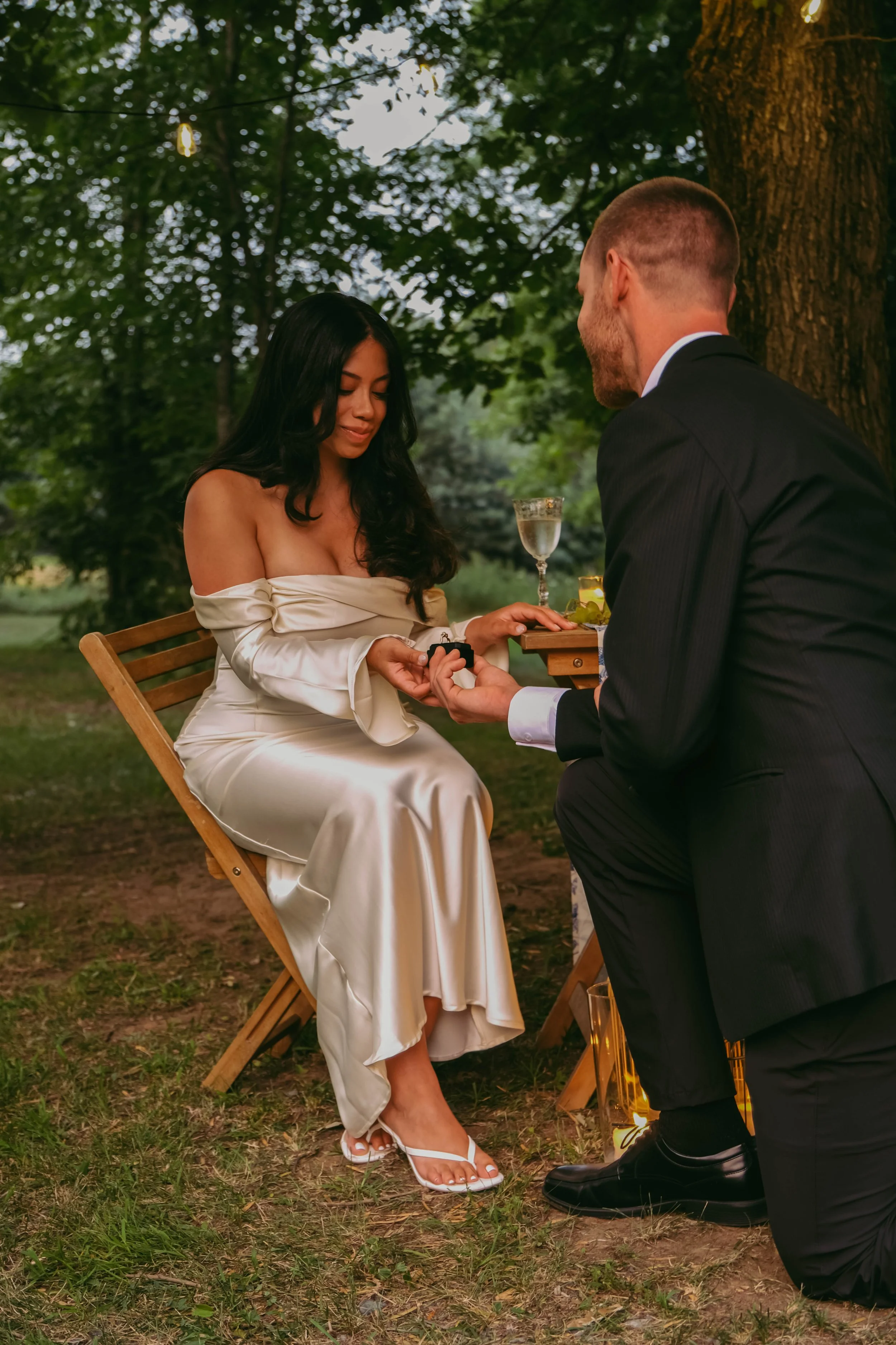 Potsdam, NY Photographer A woman in an off-the-shoulder, satin ivory dress sits on a wooden chair outdoors during sunset, holding hands with a man in a black suit who is kneeling before her, proposing marriage.