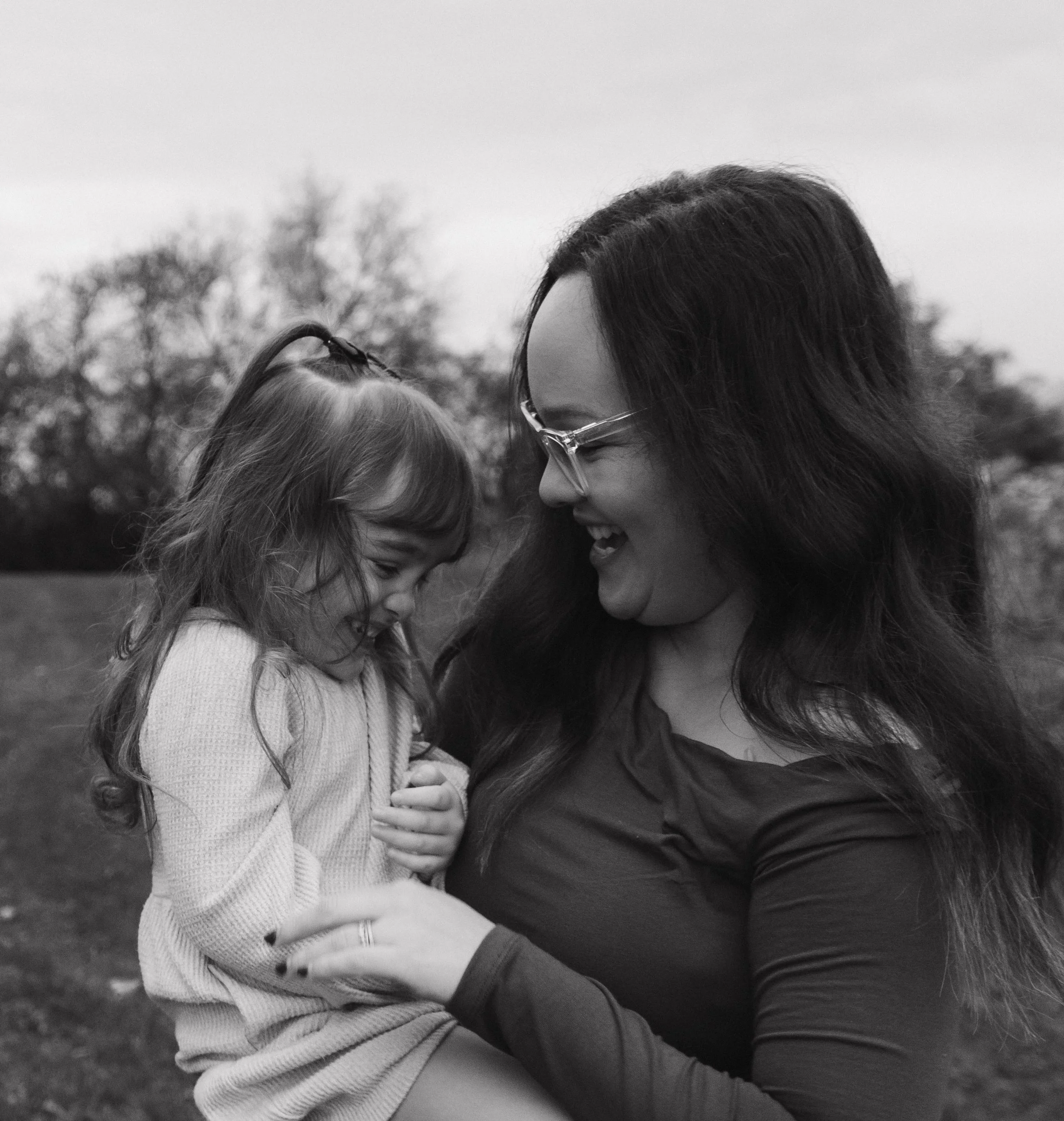 Baldwinsville, NY Photographer A woman and a young girl sharing a joyful moment outdoors, with the woman holding the girl in her arms, both smiling and looking at each other.