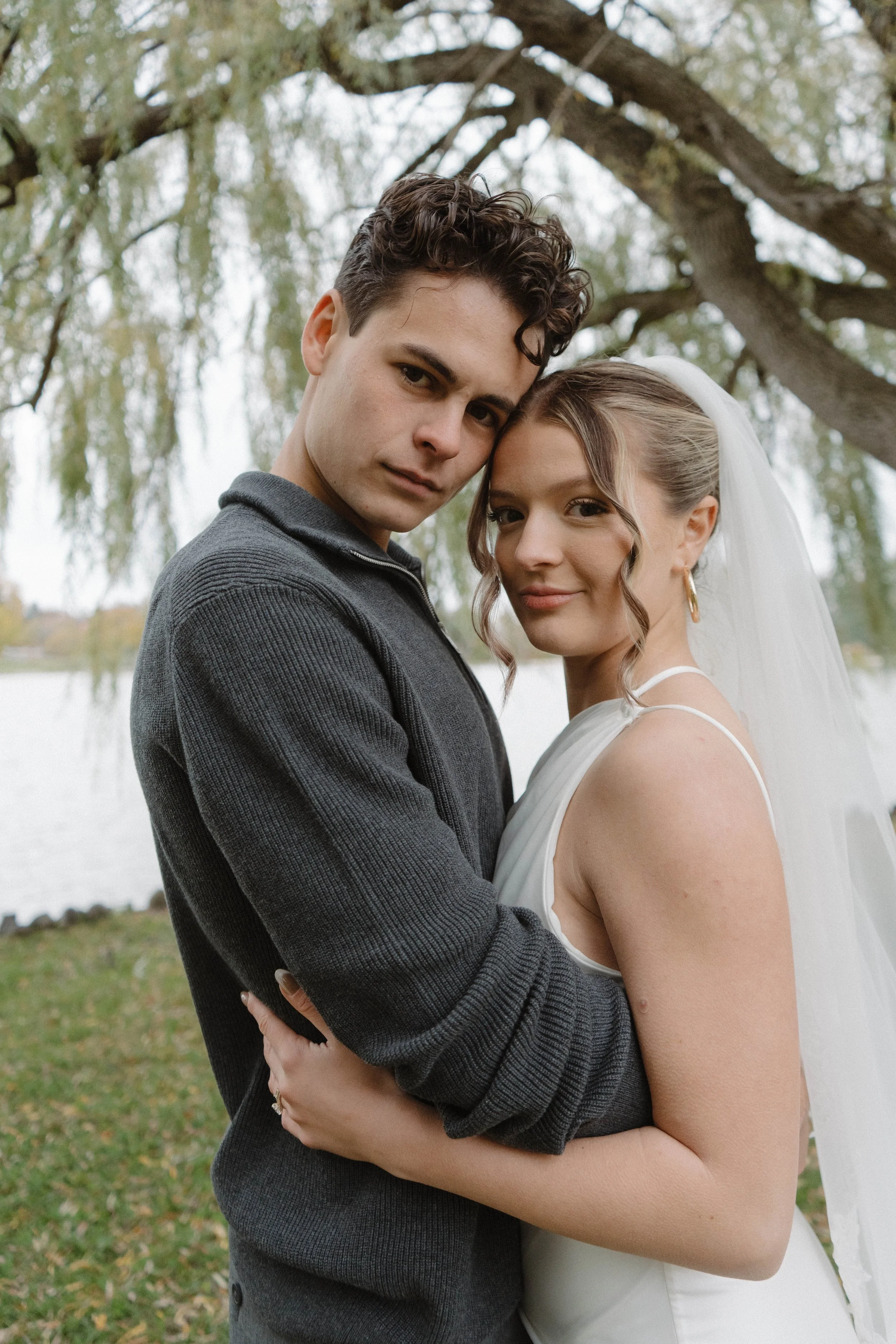 A young couple in wedding attire standing outdoors near a lake with trees and green grass in the background, embracing and looking at the camera.