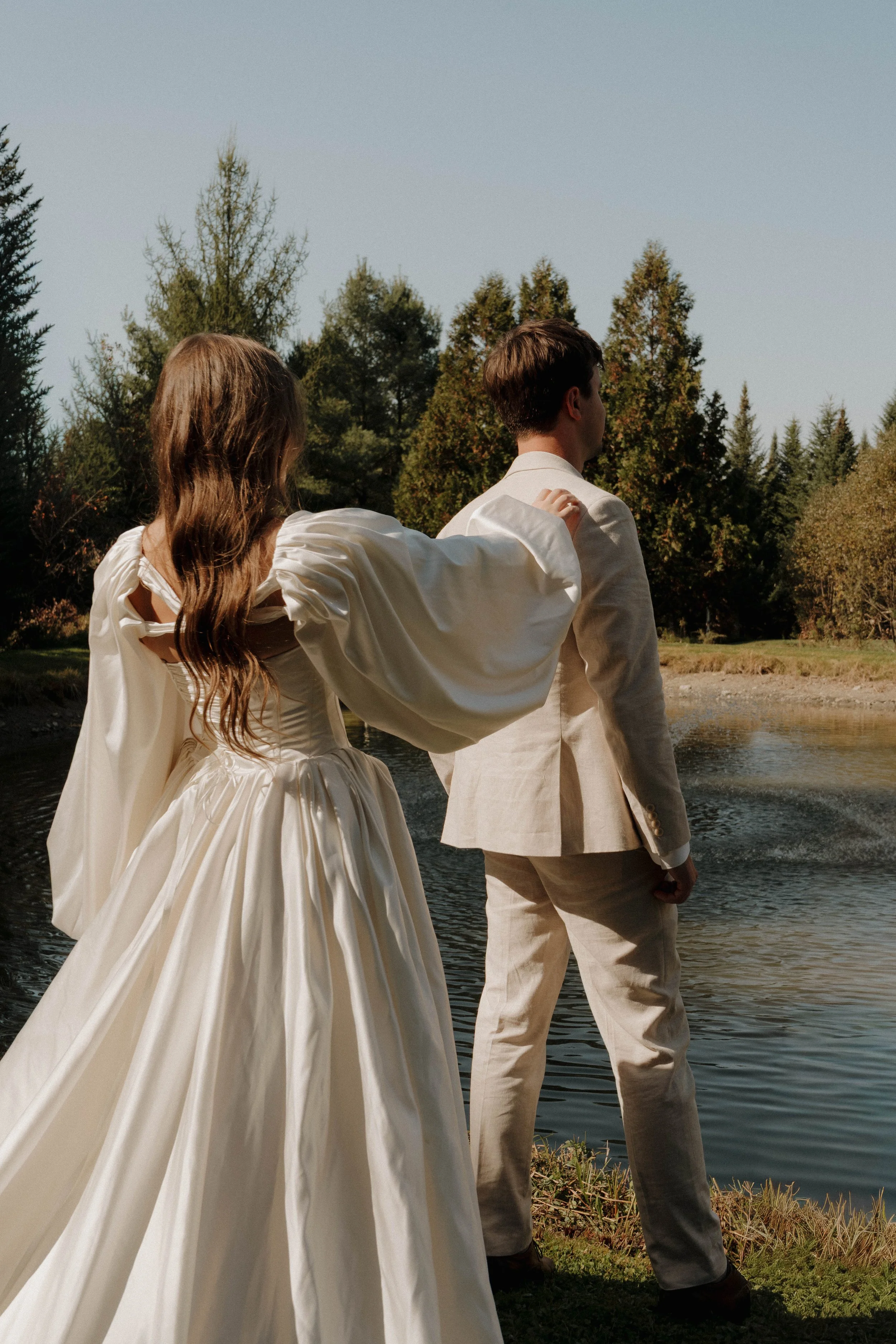 A woman in a white wedding gown with long flowing hair and a man in a cream-colored suit standing by a pond with trees in the background.