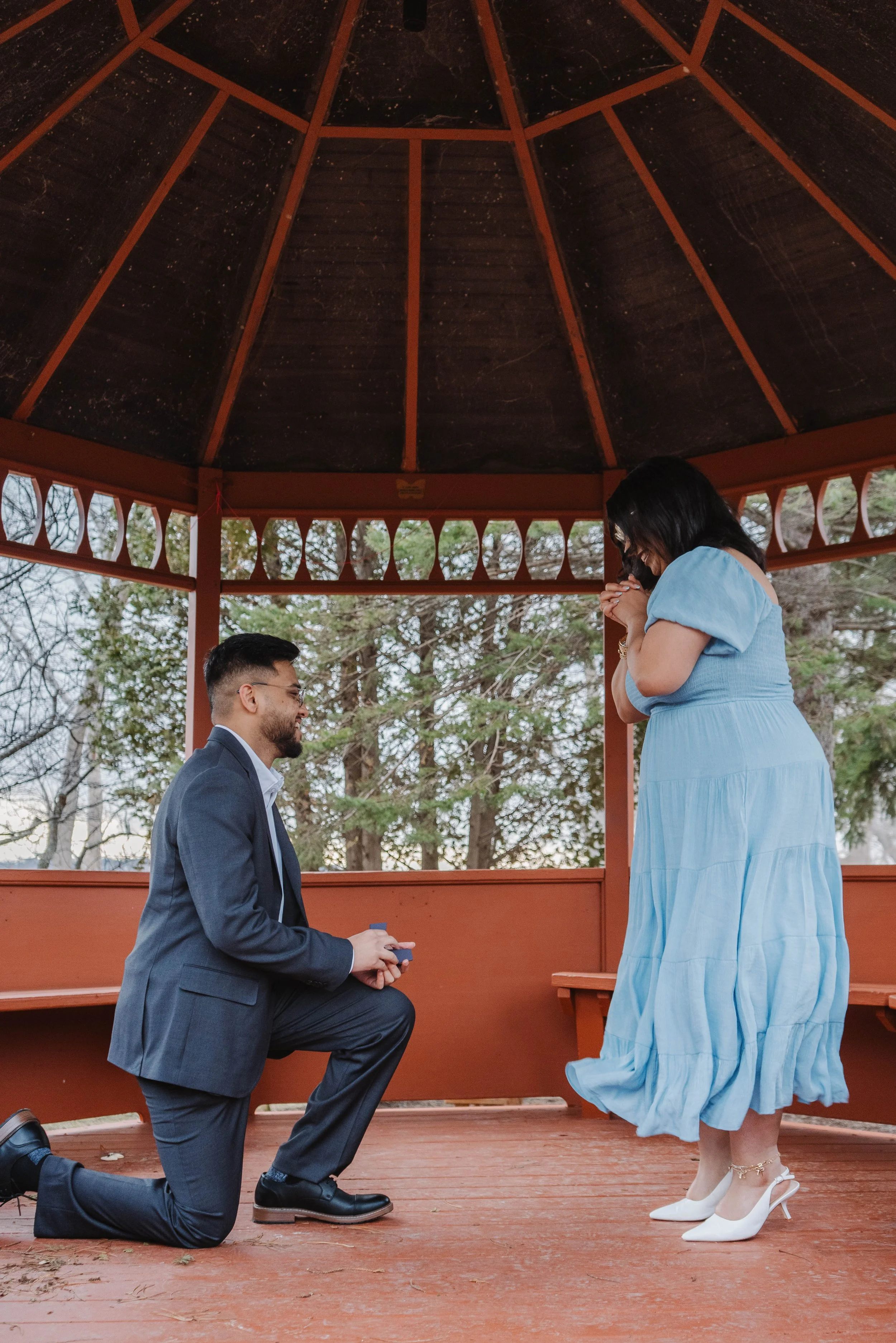 A man is kneeling on one knee holding a small box, proposing to a woman inside a wooden gazebo. The woman, dressed in a light blue dress and white heels, is visibly surprised and emotional.