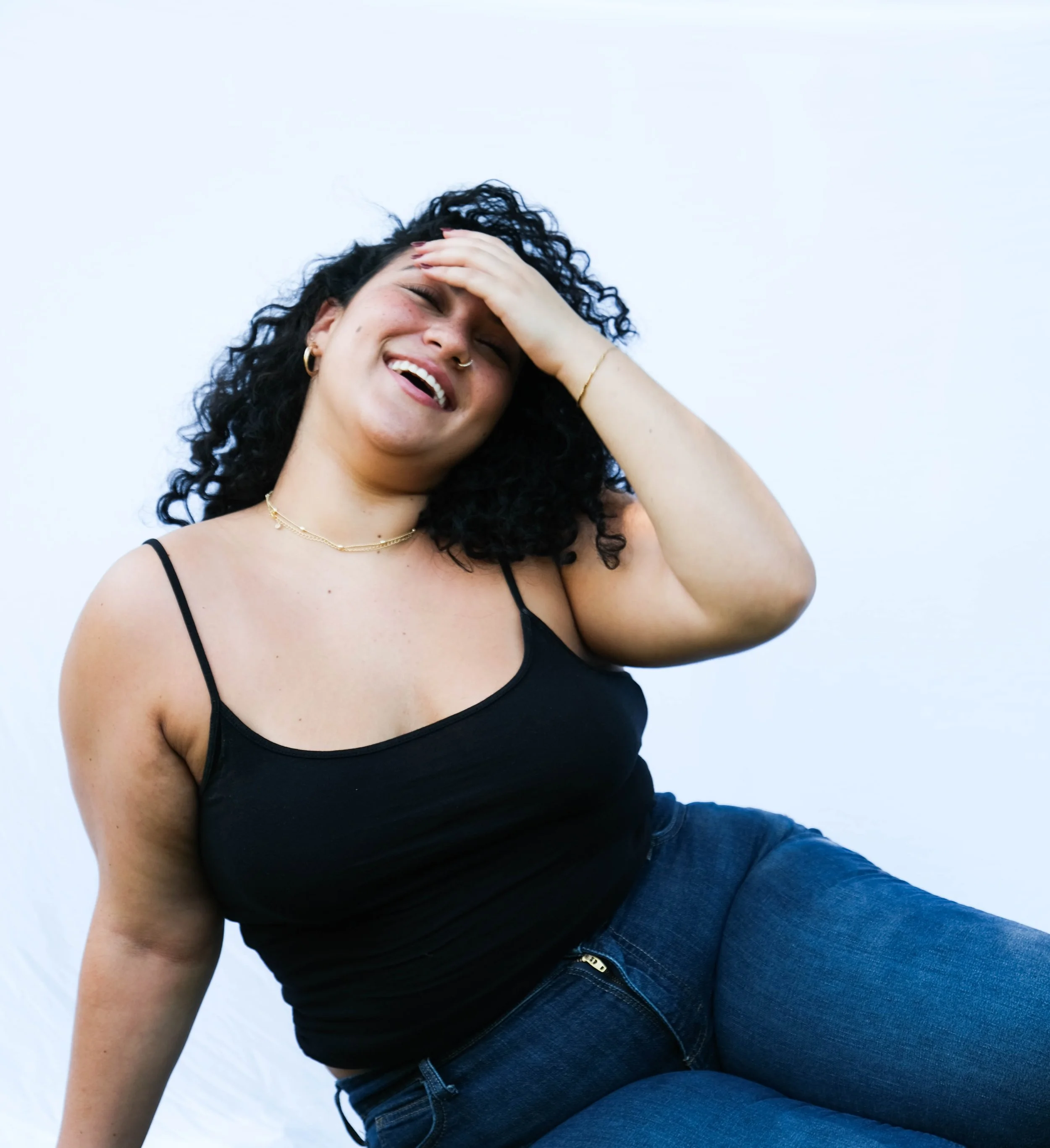 A woman with curly black hair, wearing a black spaghetti strap top and blue jeans, laughing with her hand on her forehead against a plain white background.