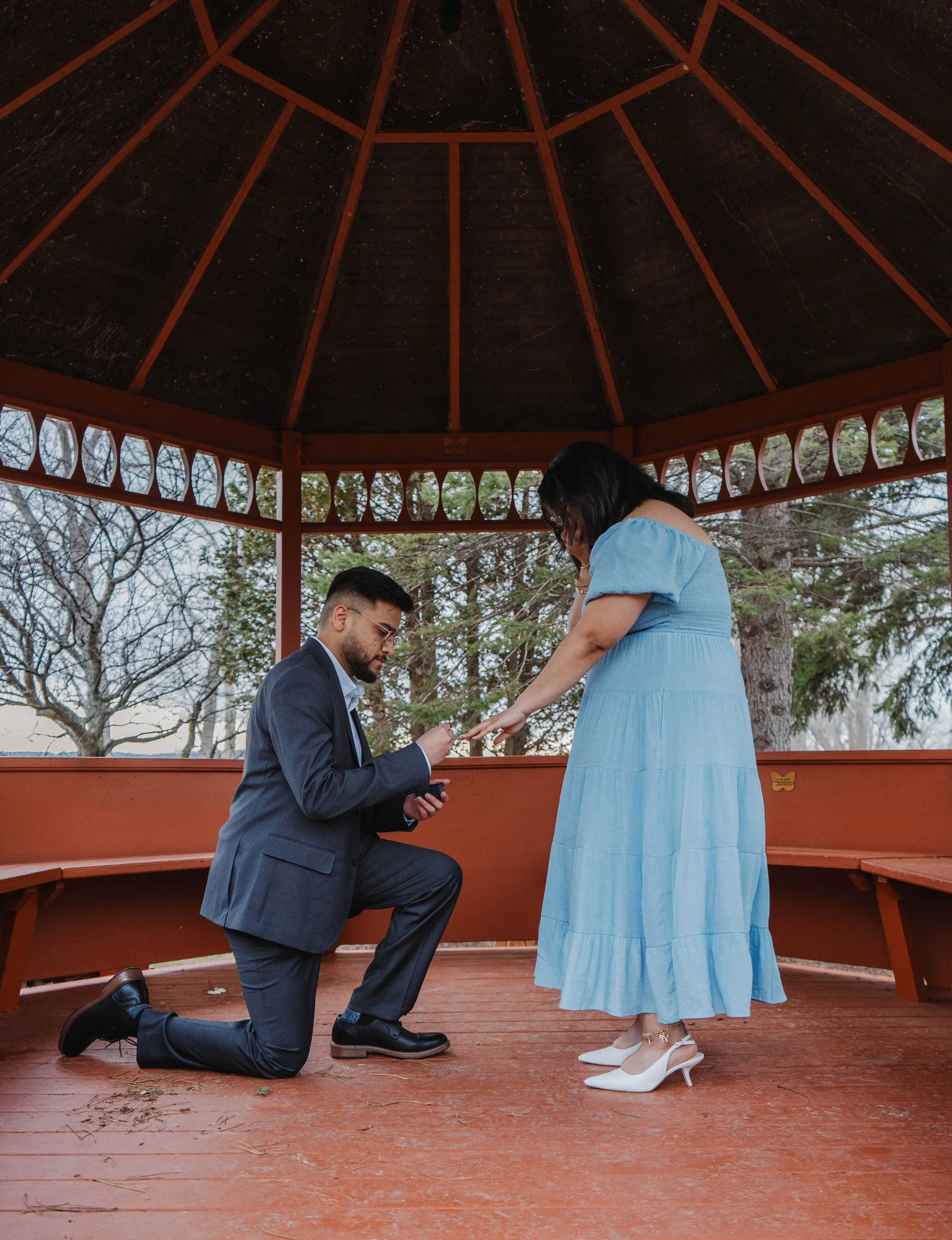 A man in a suit kneeling on one knee proposing to a woman in a blue dress and white heels, inside a wooden gazebo with trees outside in the background.