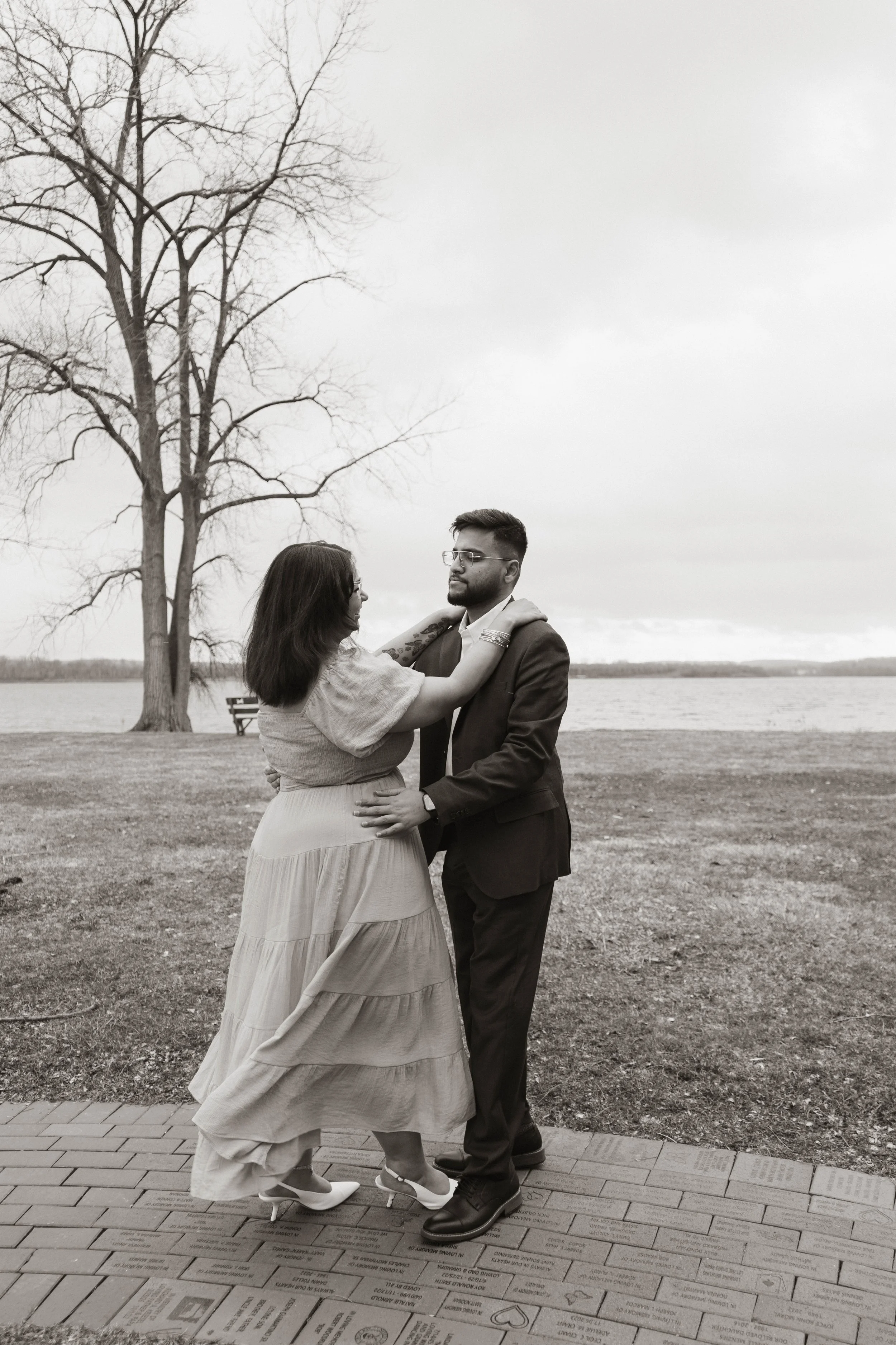 A black-and-white photo of a couple dancing outdoors near a body of water with a large, leafless tree in the background.