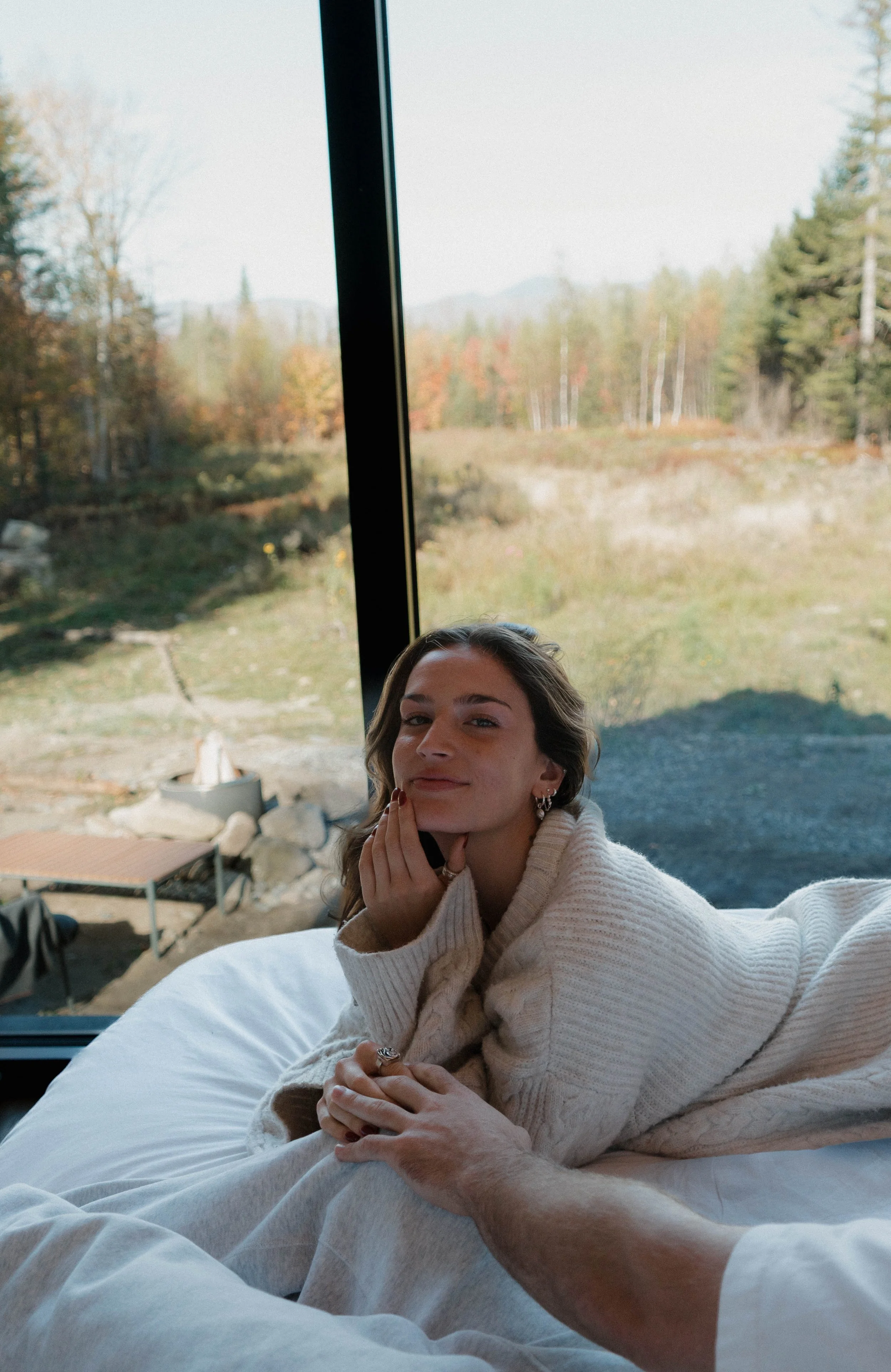 A young woman with long brown hair and earrings relaxing on a bed indoors, with a large window showing an outdoor landscape of trees and grassy area.