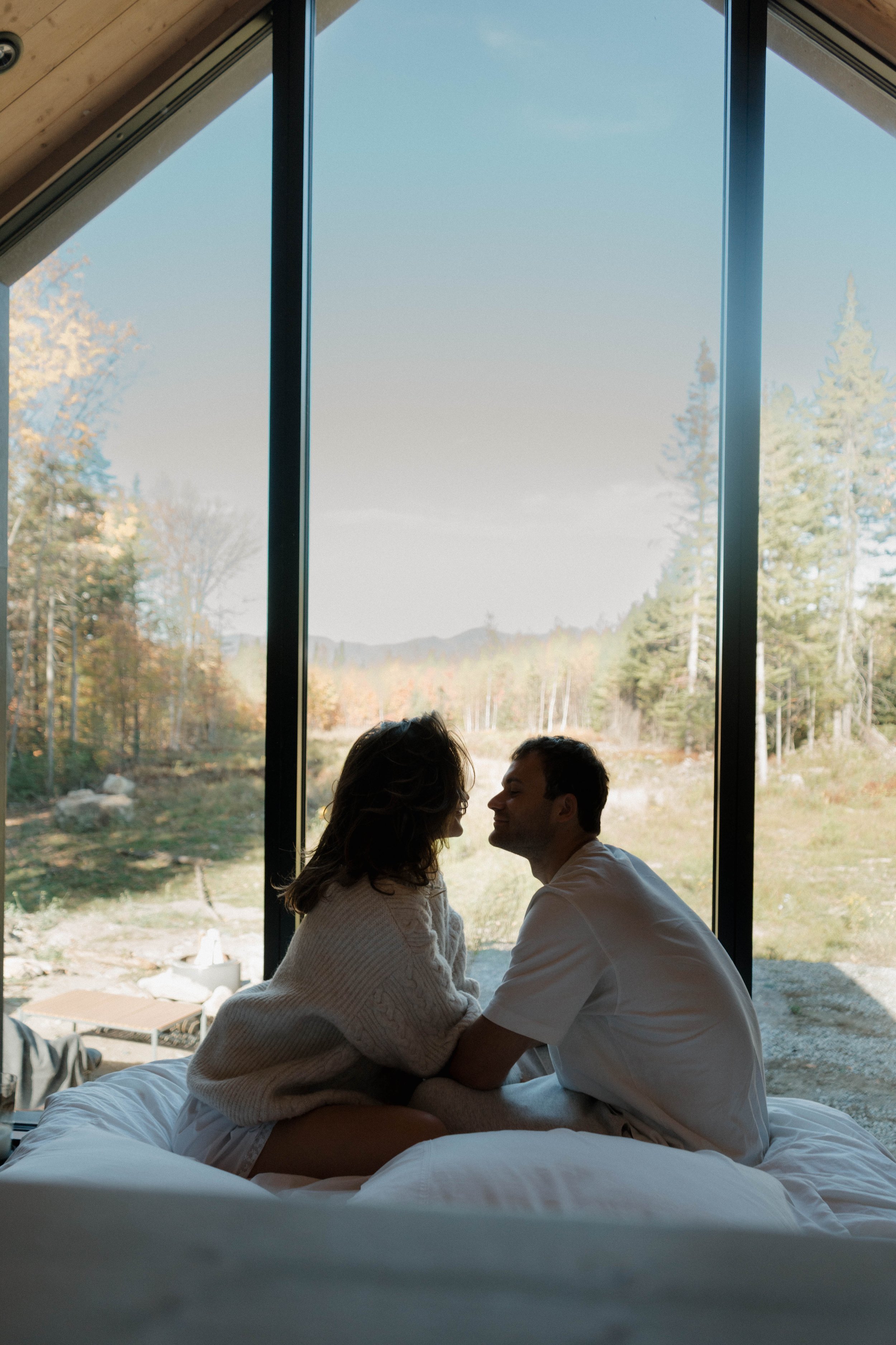 A couple sitting closely on a bed, facing each other with foreheads touching, in front of large floor-to-ceiling windows overlooking a forested landscape on a clear day.