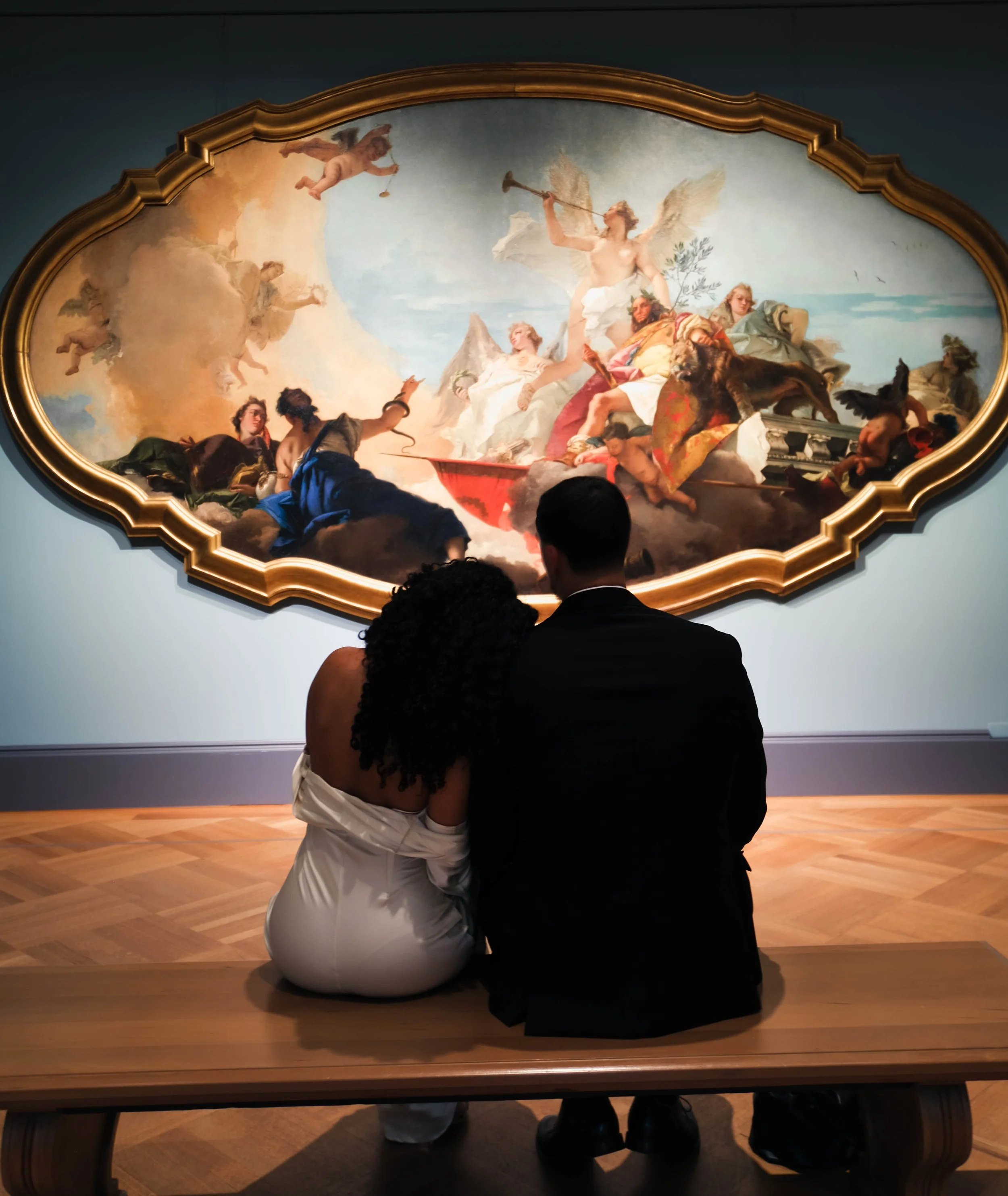The Met Engagement Shoot  A couple sitting on a bench in an art gallery, viewing a large classical painting of mythological figures and angels.