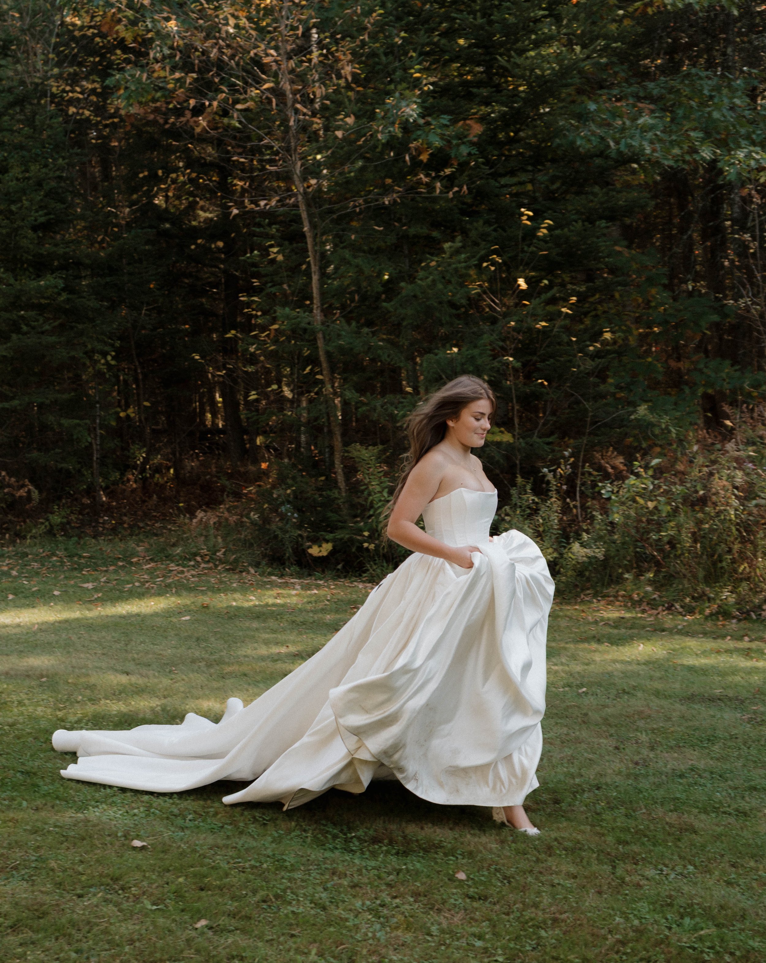Young woman in a white wedding dress walking on green grass with a forest background.