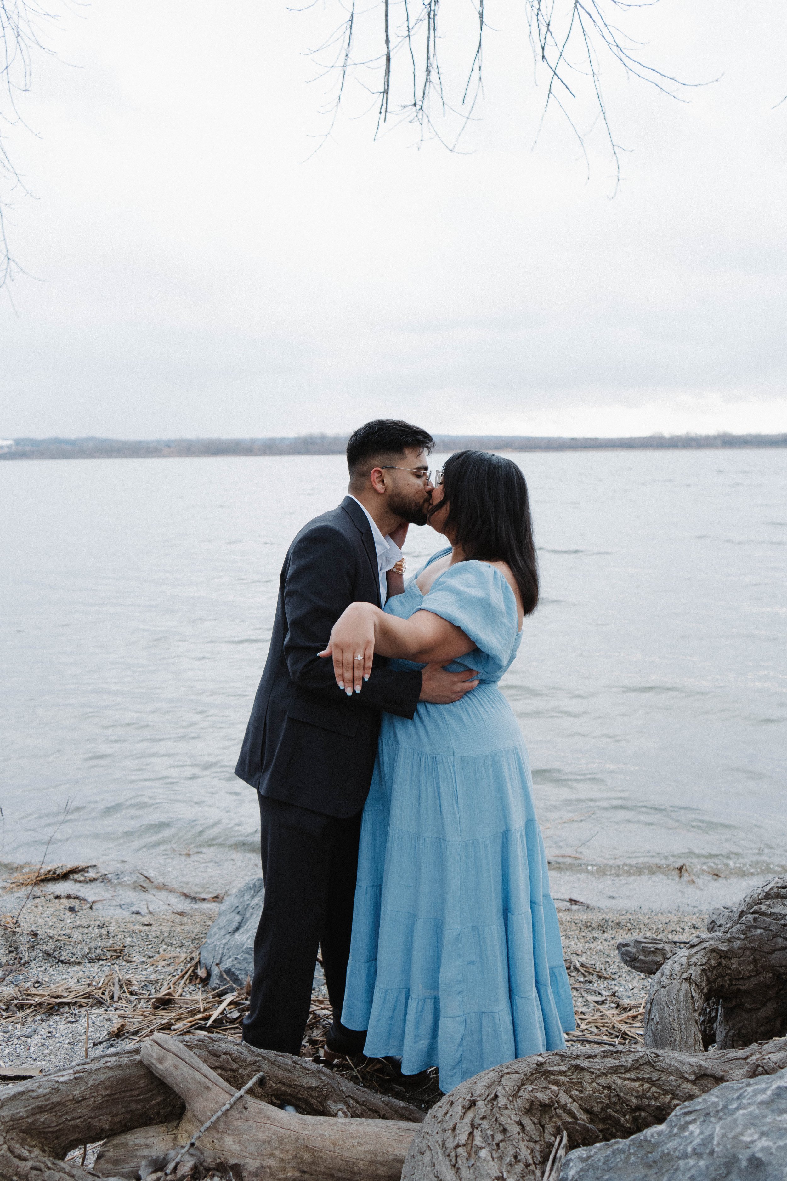 A couple kissing by a lakeside, the woman is in a light blue dress and the man is in a dark suit.