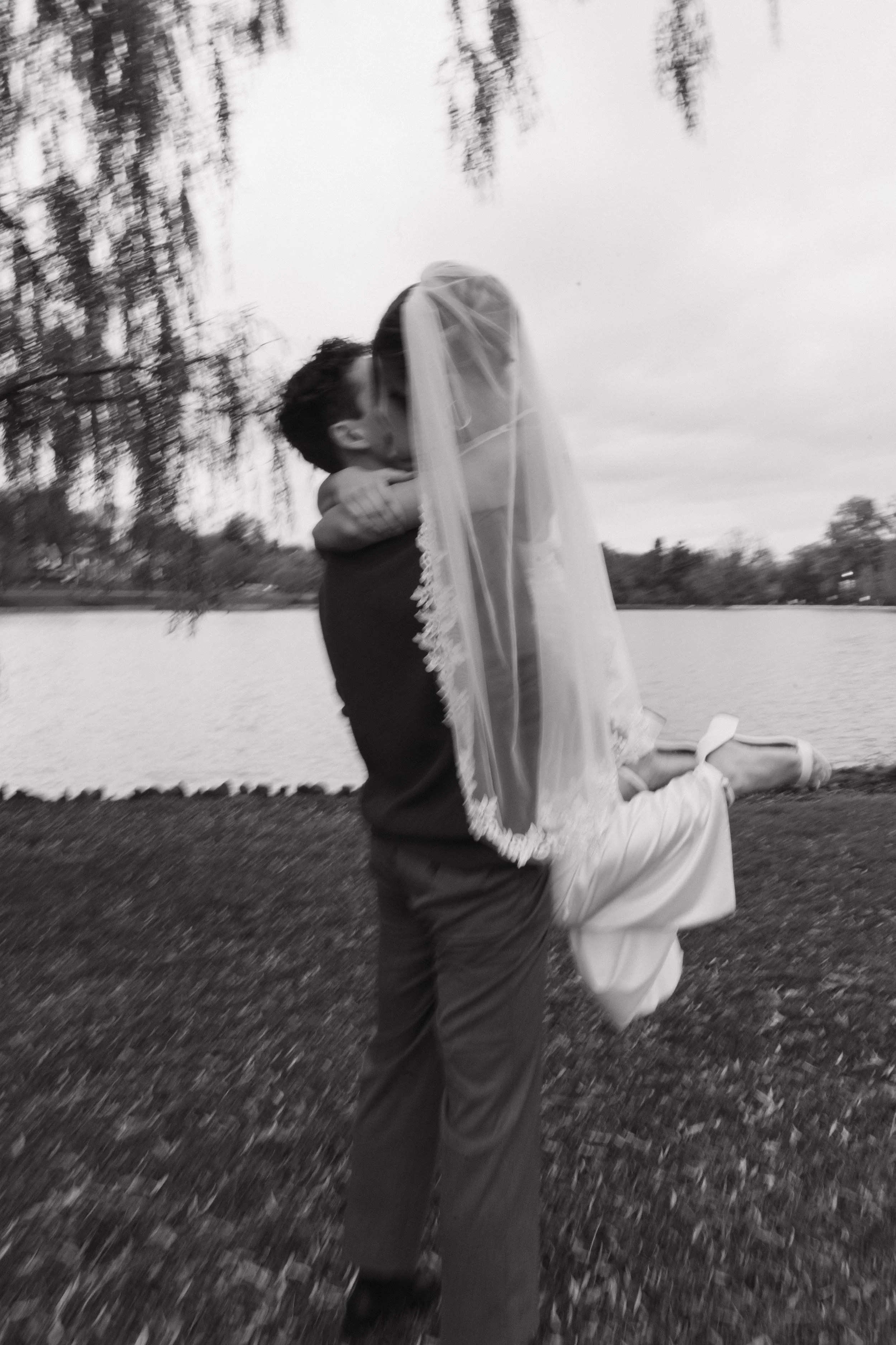 A black and white photo of a man lifting a woman in a wedding dress in front of a lake, with trees in the background.