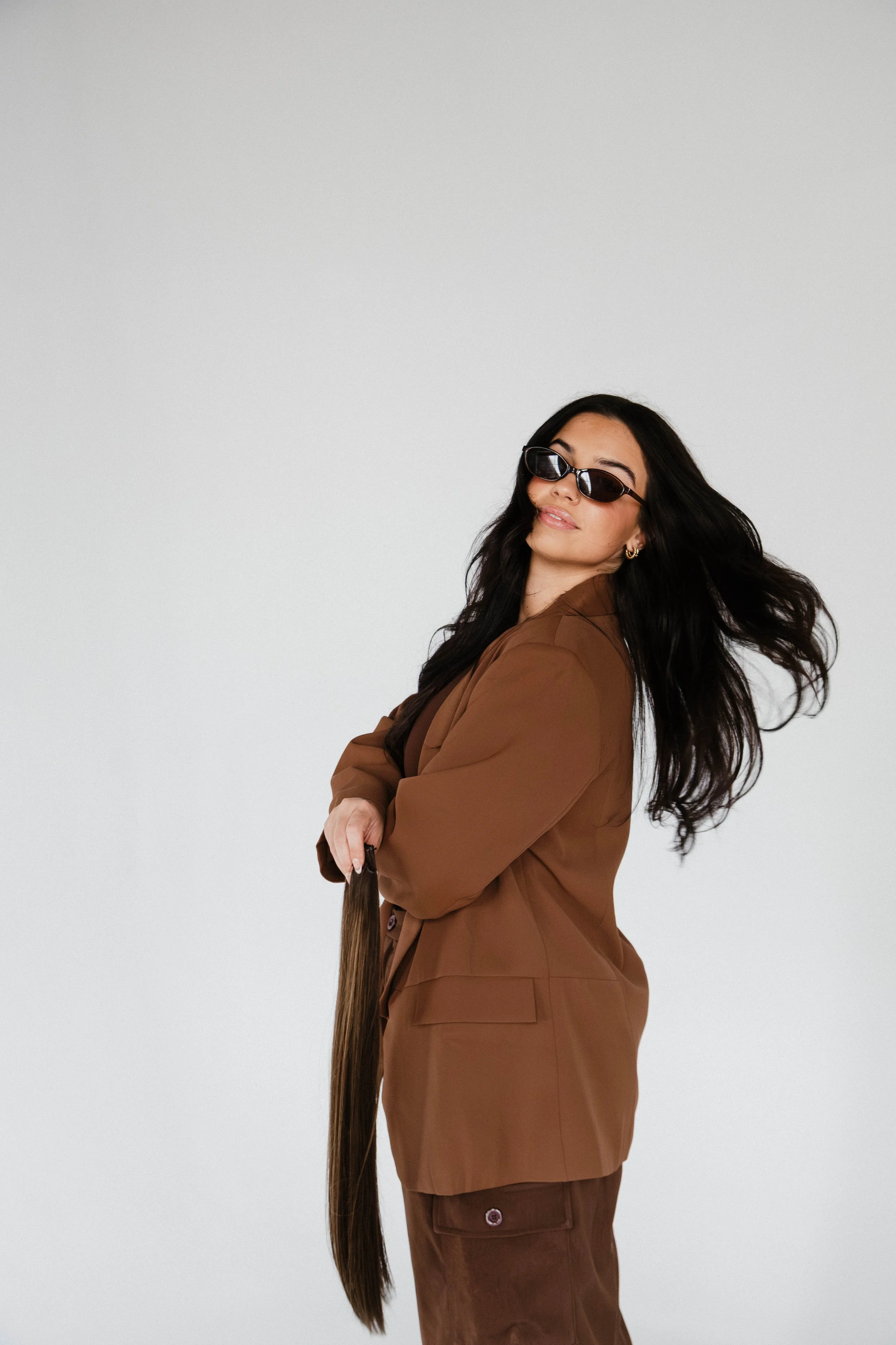 Syracuse, NY Branding photos, A woman with long dark hair wearing sunglasses and a brown suit, holding a long hair extension, poses against a plain white background.