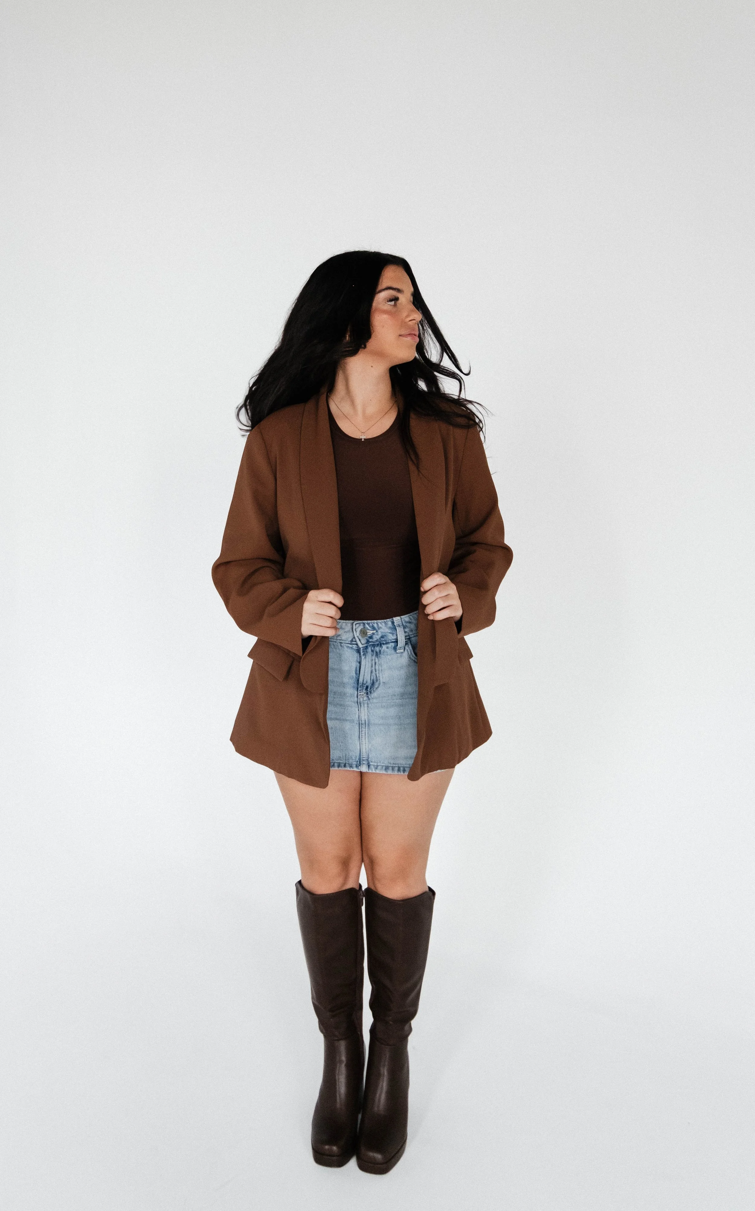 A woman with long dark hair wearing a brown blazer, black top, denim skirt, and brown knee-high boots standing against a plain white background.