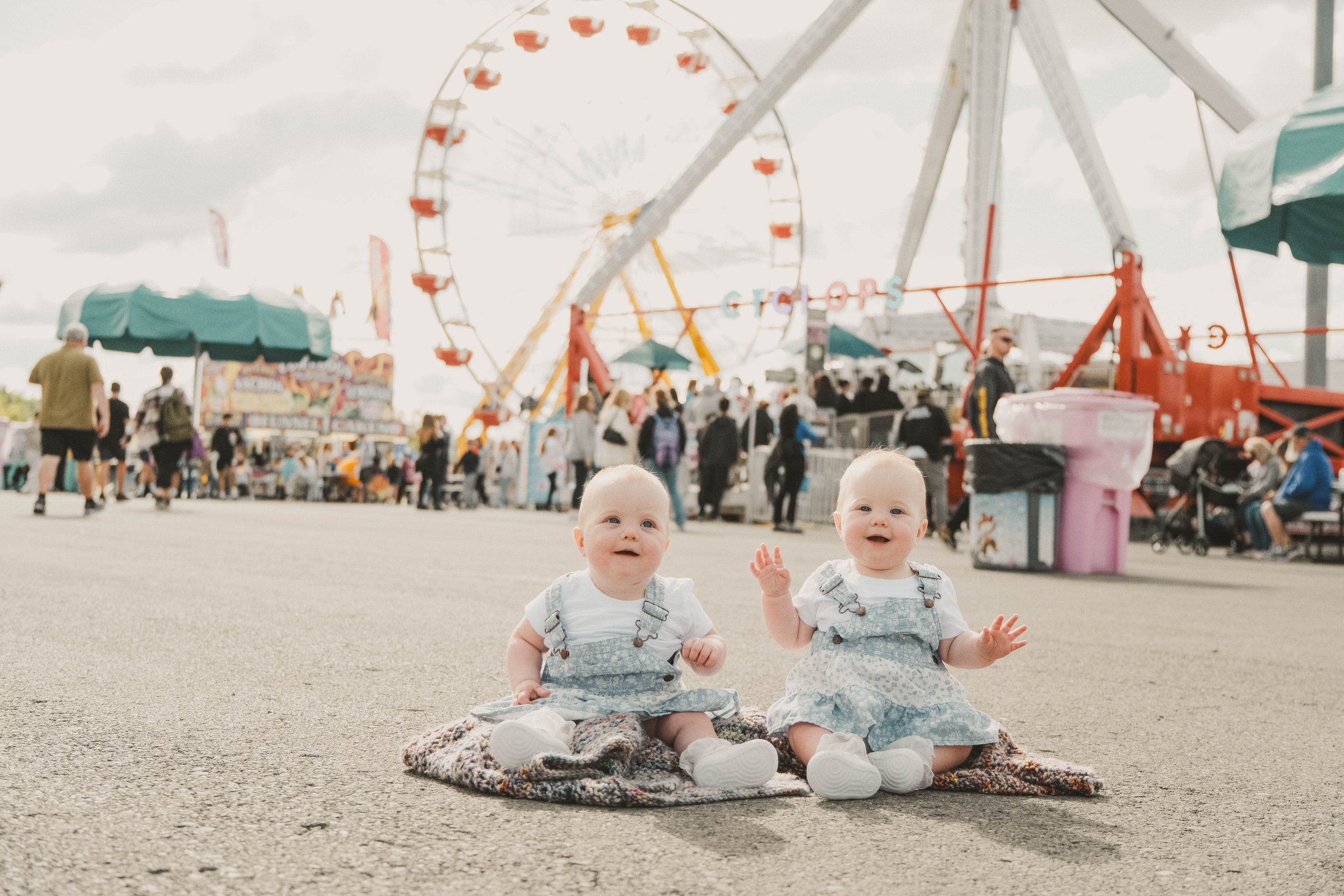 NYS Fair Minis, Syracuse, NY, Two babies sitting on a blanket at a carnival with a Ferris wheel and rides in the background.