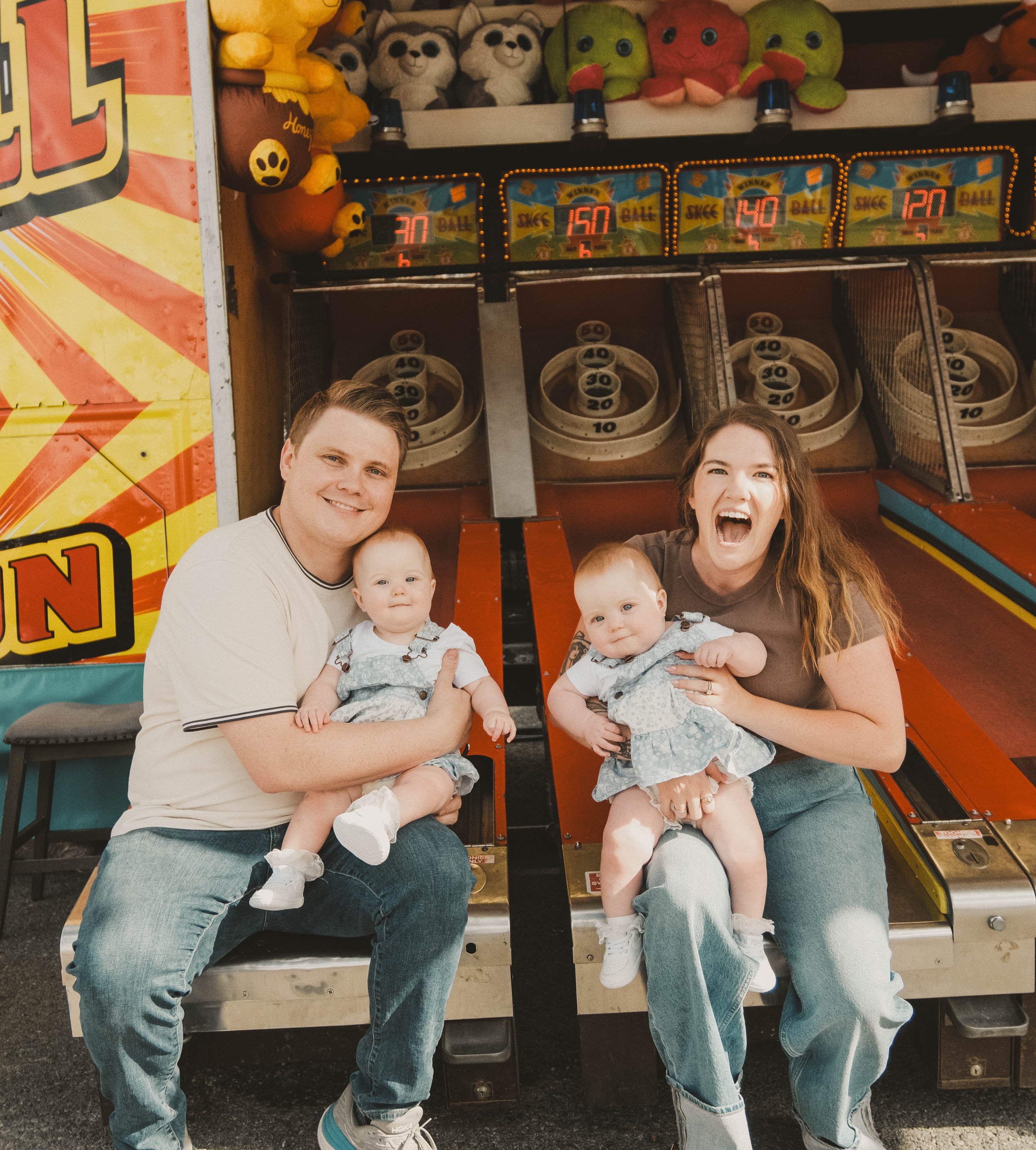 NYS Fair Minis, Syracuse, NY, A family at an arcade with two adults and two babies sitting on a bench, holding the babies, with a skee-ball game in the background.