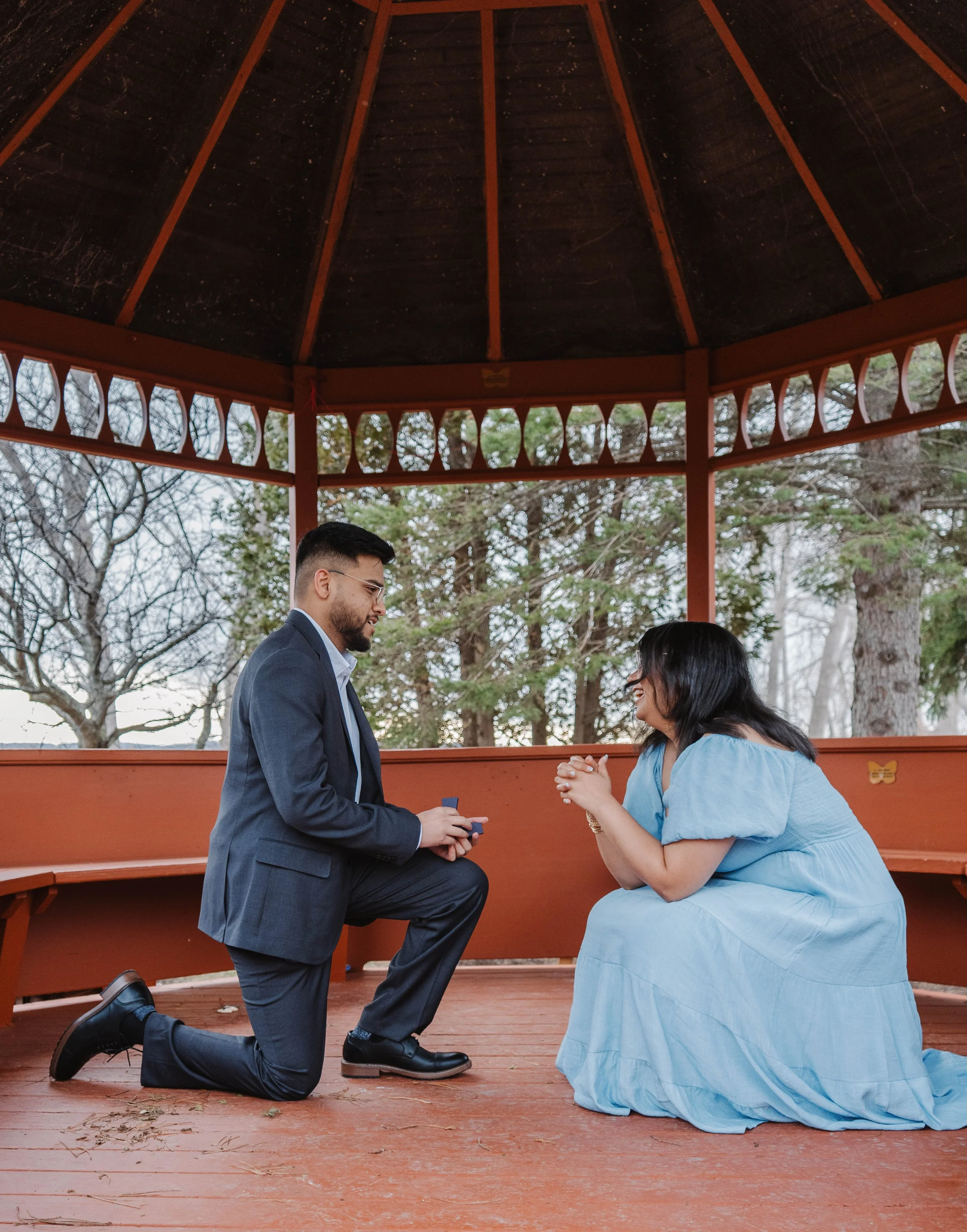 A man dressed in a suit is kneeling on one knee, holding a ring box, proposing to a woman in a blue dress who is sitting on her knees with hands clasped together, inside a round, open pavilion surrounded by trees.