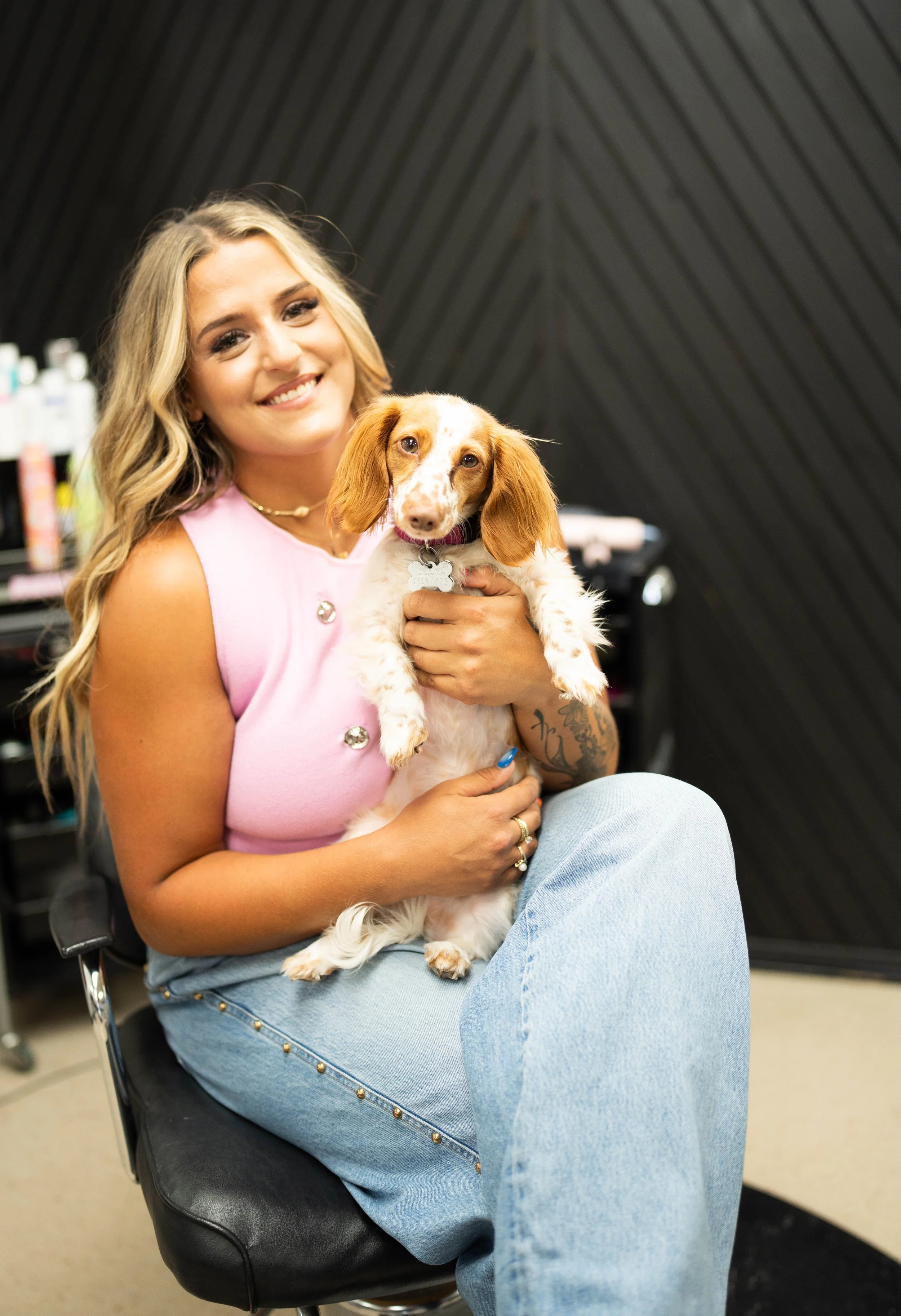 A woman with long, wavy blonde hair, smiling and wearing a pink sleeveless top, sitting in a black chair and holding a small brown and white dog with a pink collar. The background is dark with shelves and equipment.