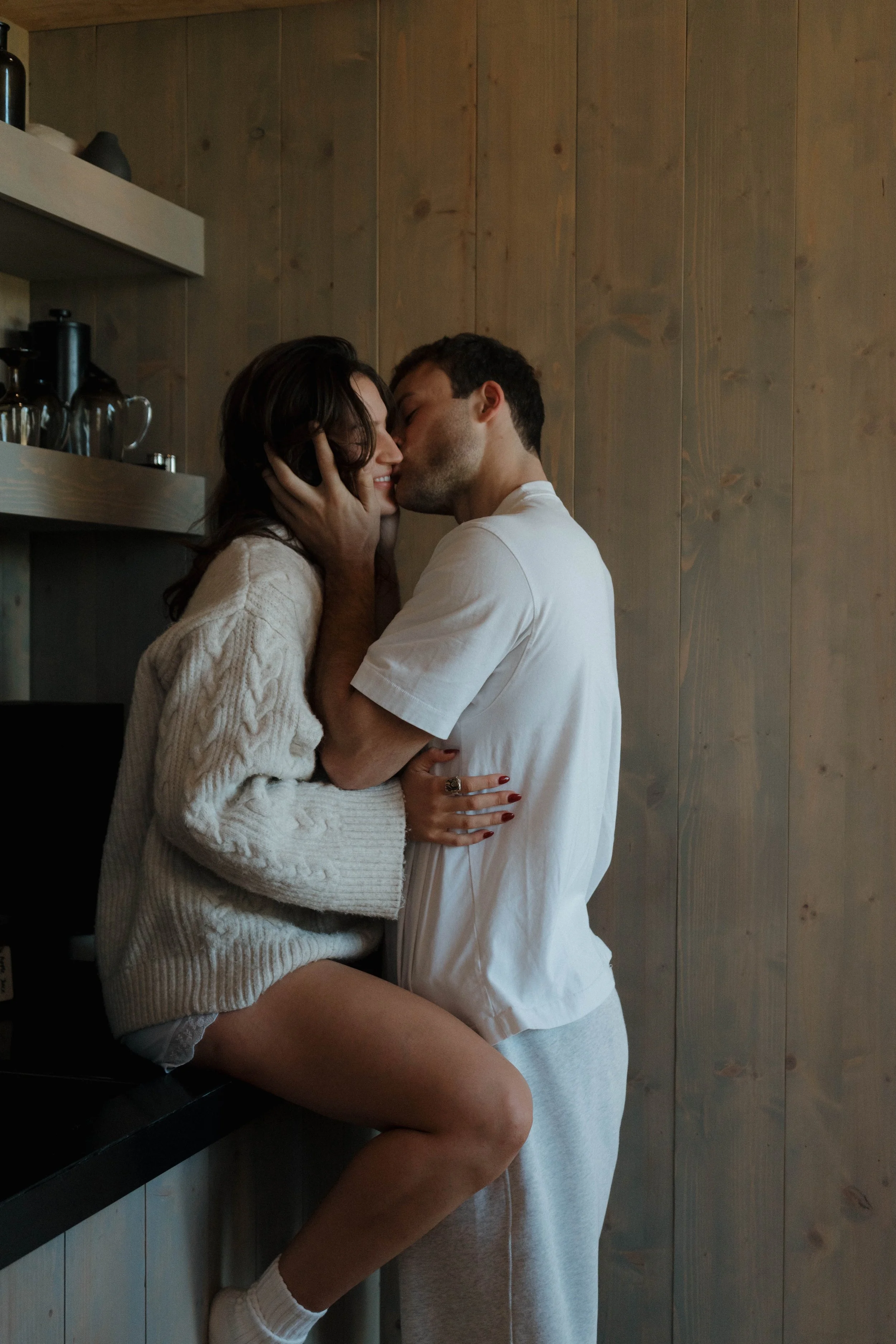 A man and woman share a kiss in a cozy, wooden-paneled kitchen. The woman, sitting on the counter, wears a cream cable knit sweater, and the man is in a white t-shirt and grey sweatpants. They are close, with the woman smiling and holding the man's face, while the man gently holds her face.