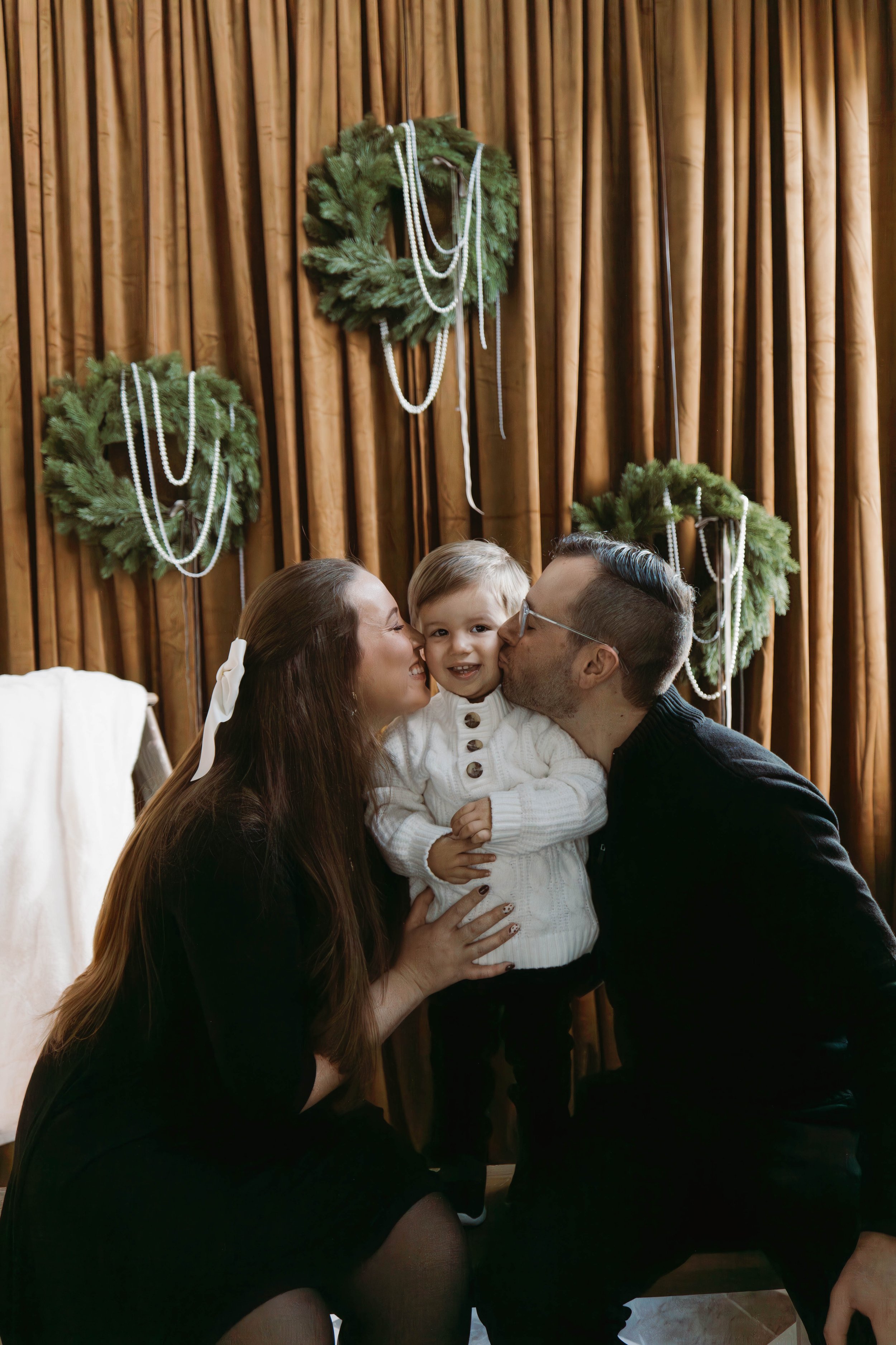 Pose Studio Photographer, A family of three at a holiday gathering, with the child and father giving kisses to the mother, who is smiling. The background features brown curtains and three green wreaths decorated with pearl garlands.
