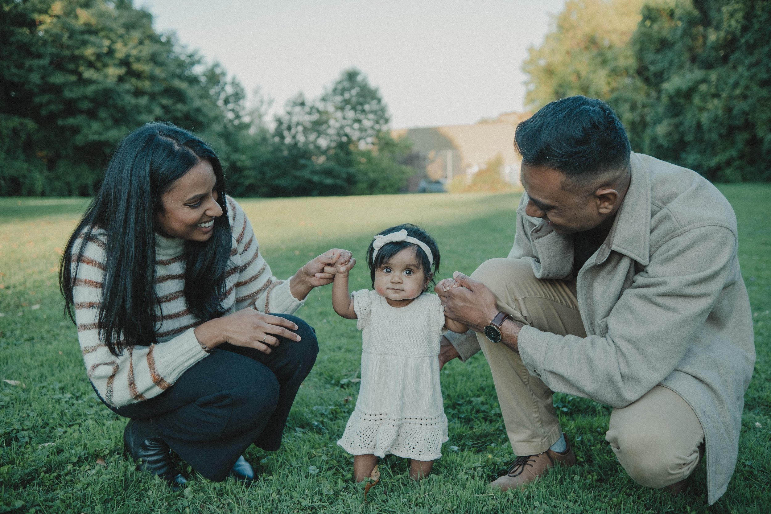 Burnet Park, Syracuse NY A happy family of three enjoying time outdoors in a park with green grass and trees in the background. The mother, father, and their young daughter are smiling and holding hands.