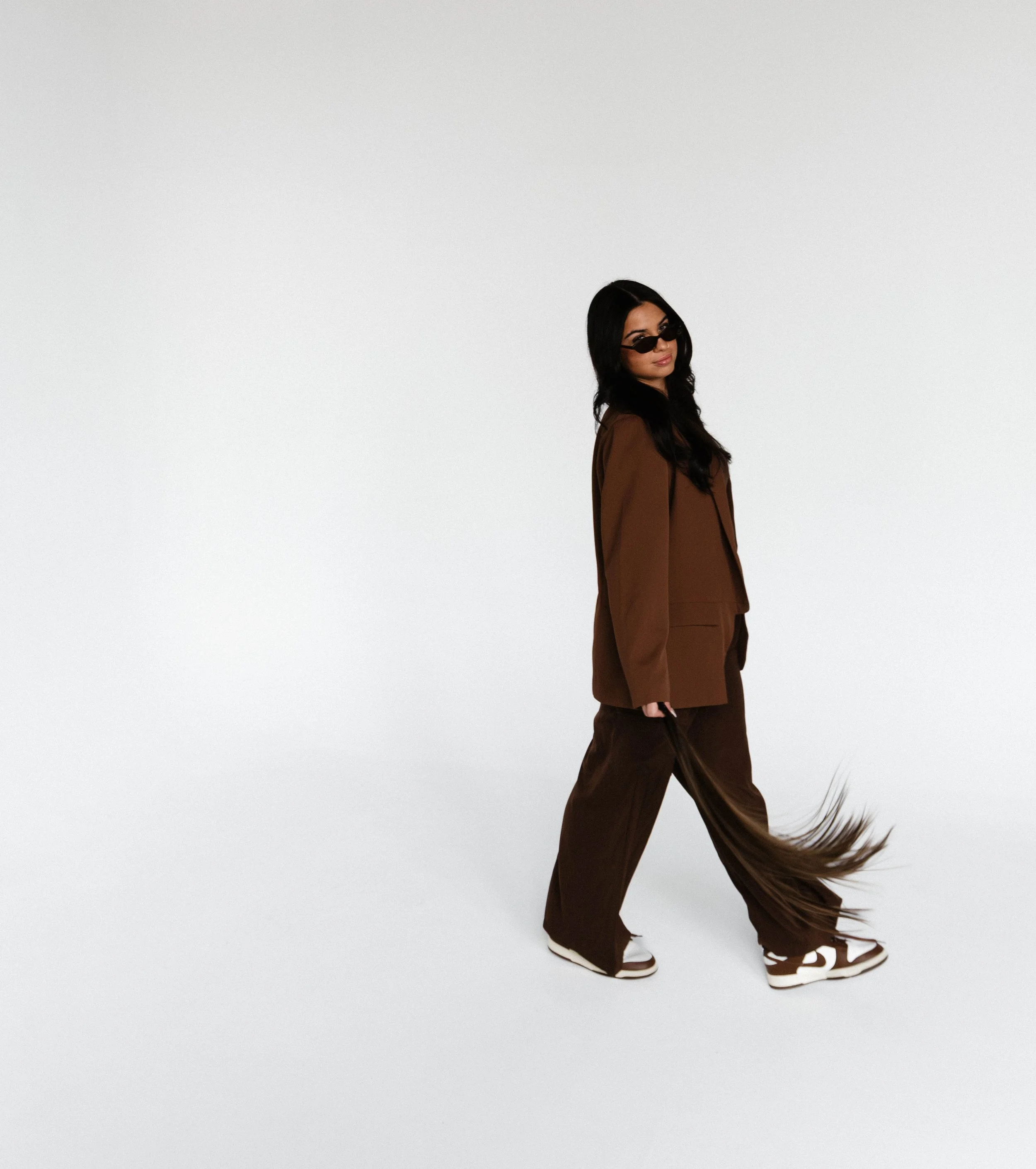 Syracuse, NY Branding photos, A woman with long dark hair, wearing sunglasses, a brown suit, and sneakers, stands against a plain white background, with her hair and pants flowing as if caught in motion.