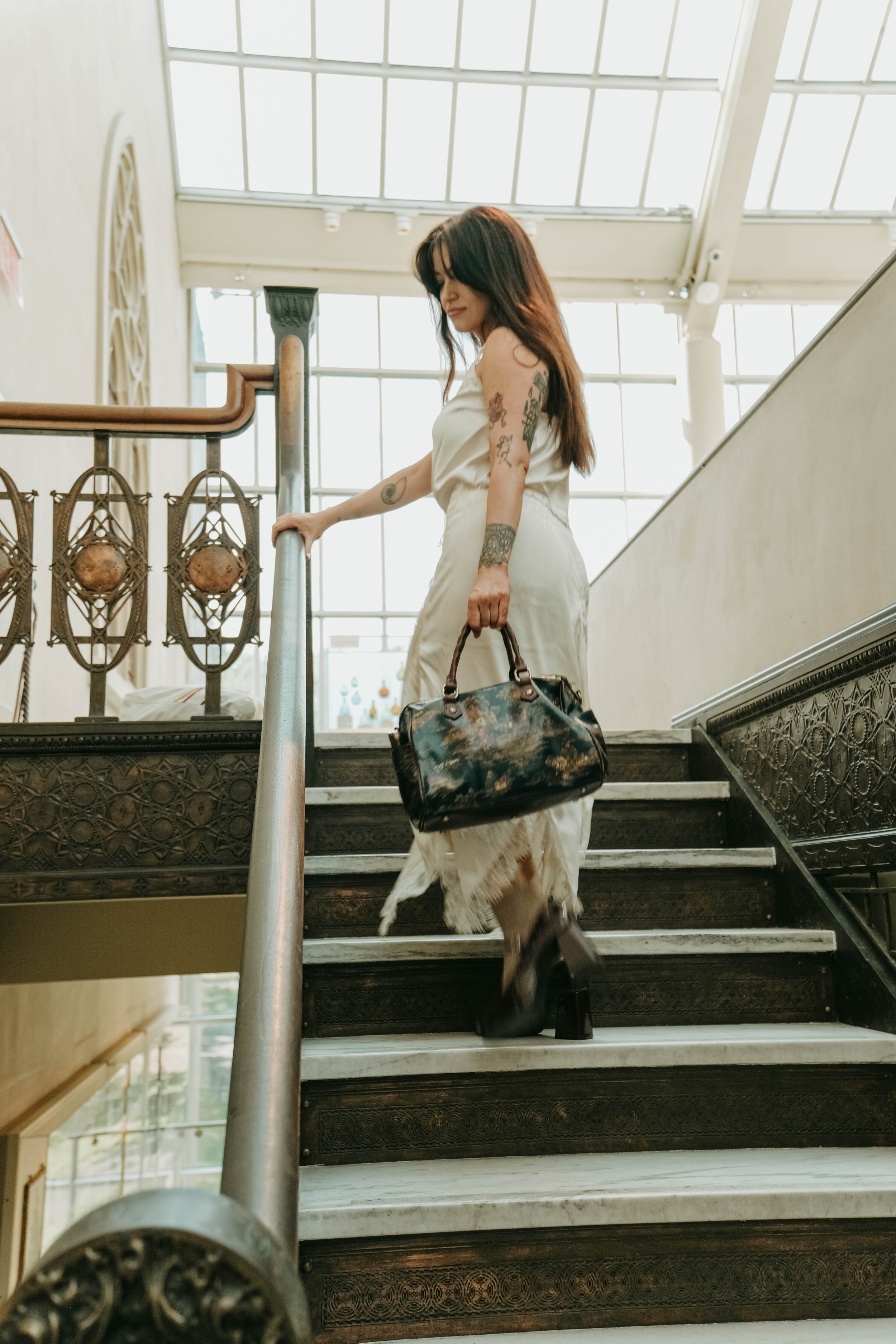The Met Museum, A woman with tattoos wearing a white dress and heels, descending a staircase inside a building with large windows, carrying a dark handbag.