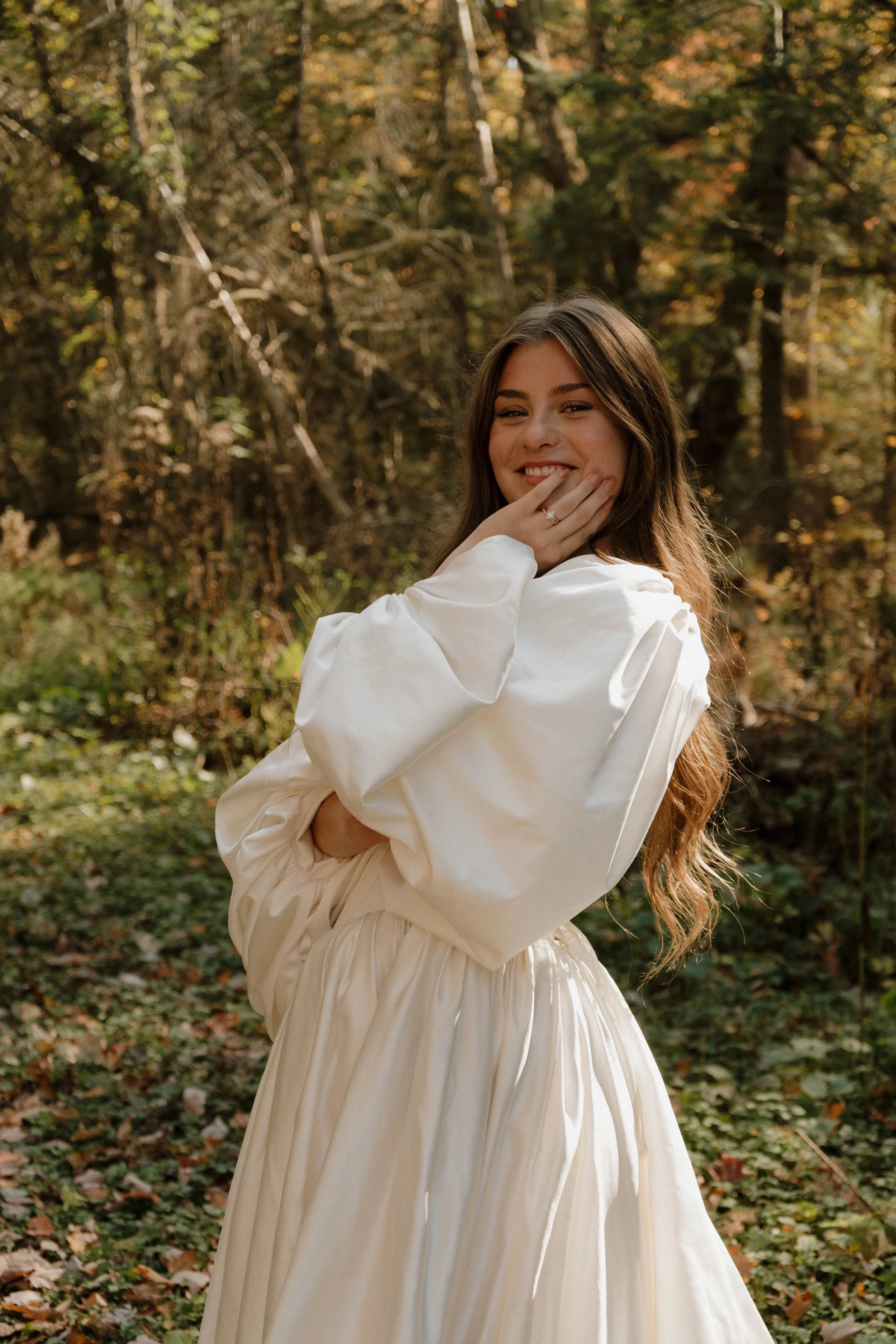 Irasburg, Vermont A young woman with long brown hair, wearing a white dress with puffed sleeves, standing in a forest with autumn foliage, smiling and touching her face with her hand.