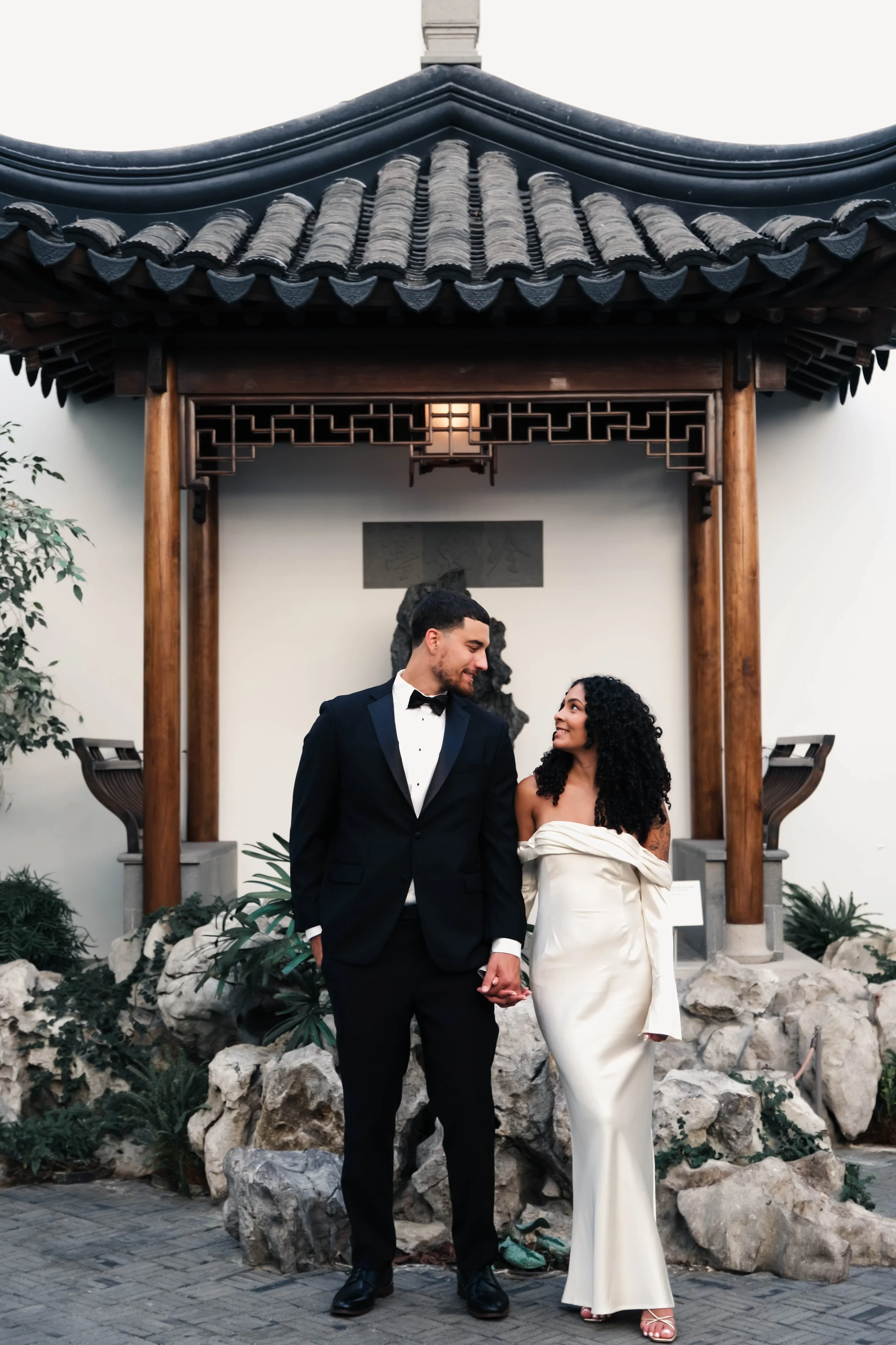 The Met Engagement Shoot  A couple dressed in formal attire, holding hands and smiling at each other, standing outdoors in front of traditional Asian architecture with rocks and plants around.