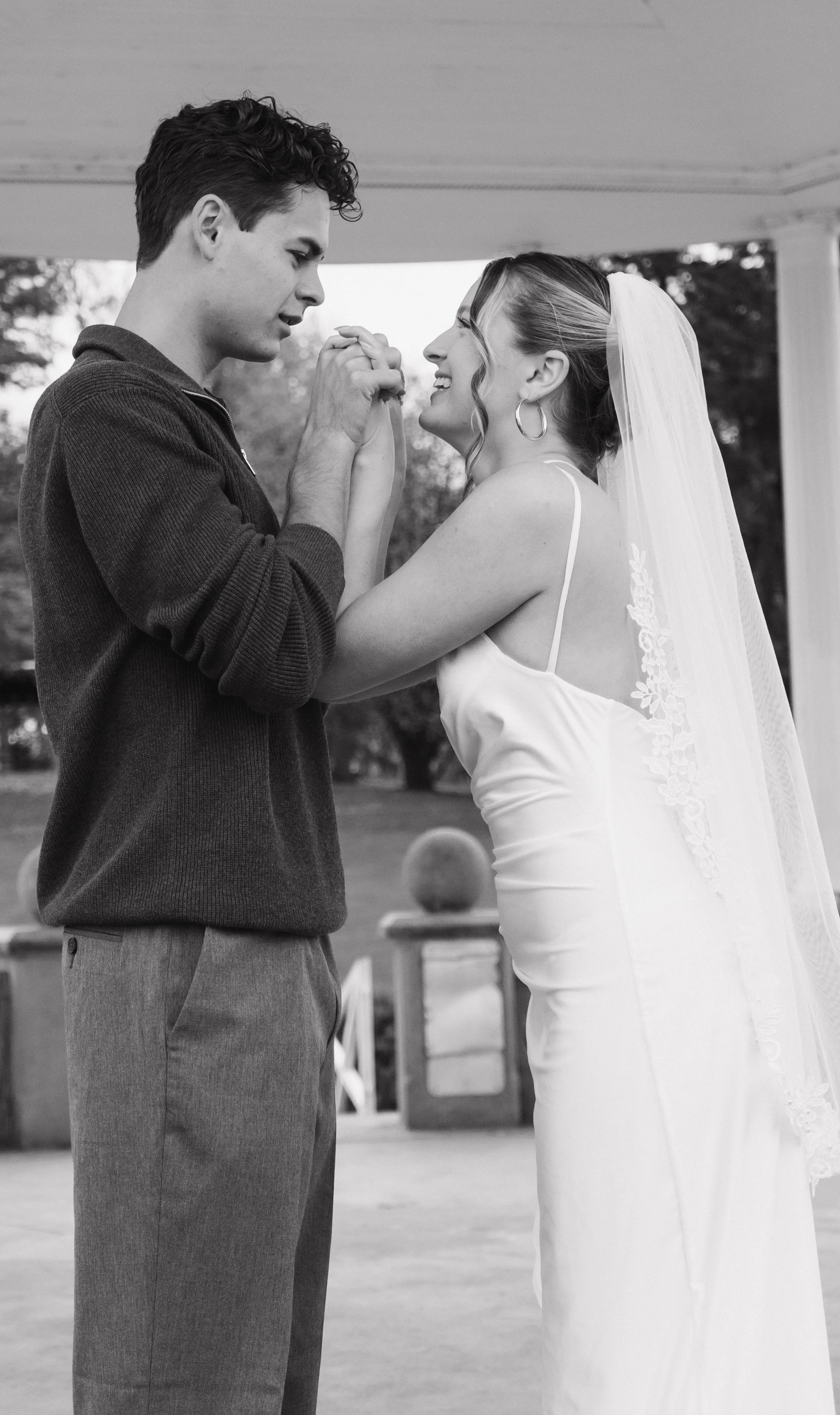 A bride and groom share a moment outside, holding hands and smiling at each other. The bride is wearing a wedding dress and veil, and the groom is dressed in casual clothes.
