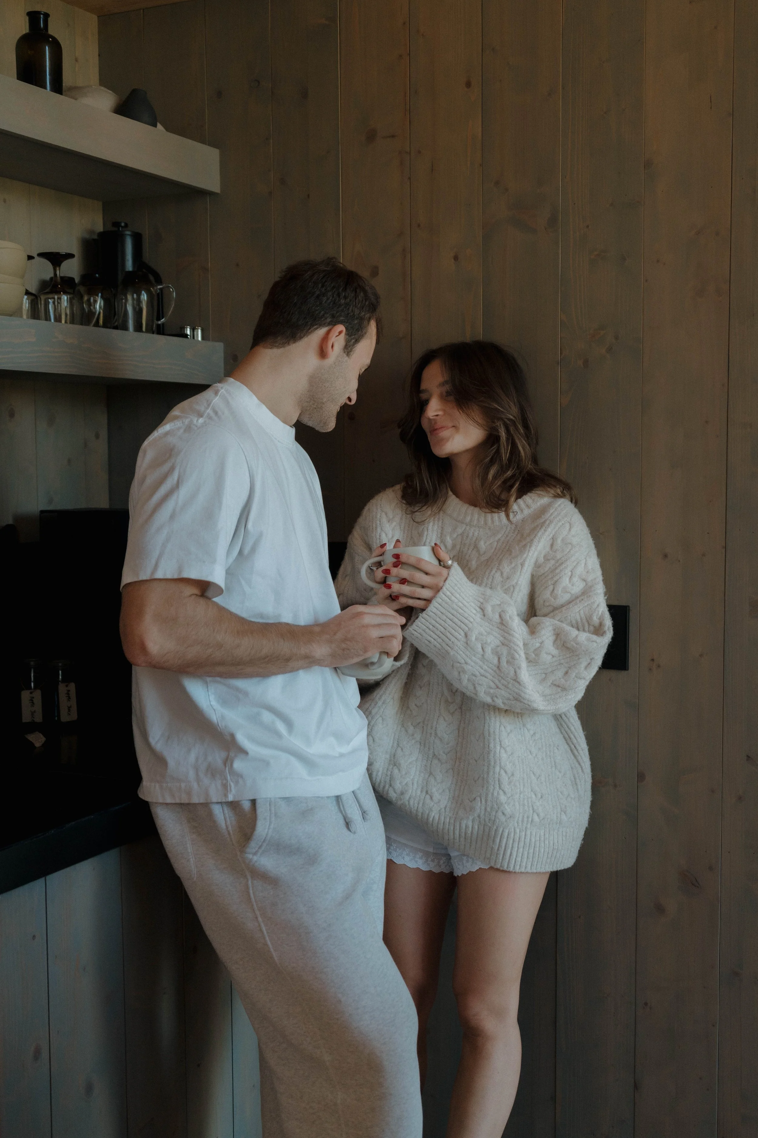 A man and woman standing in a cozy kitchen, sharing a moment with the woman holding a mug and both leaning towards each other.