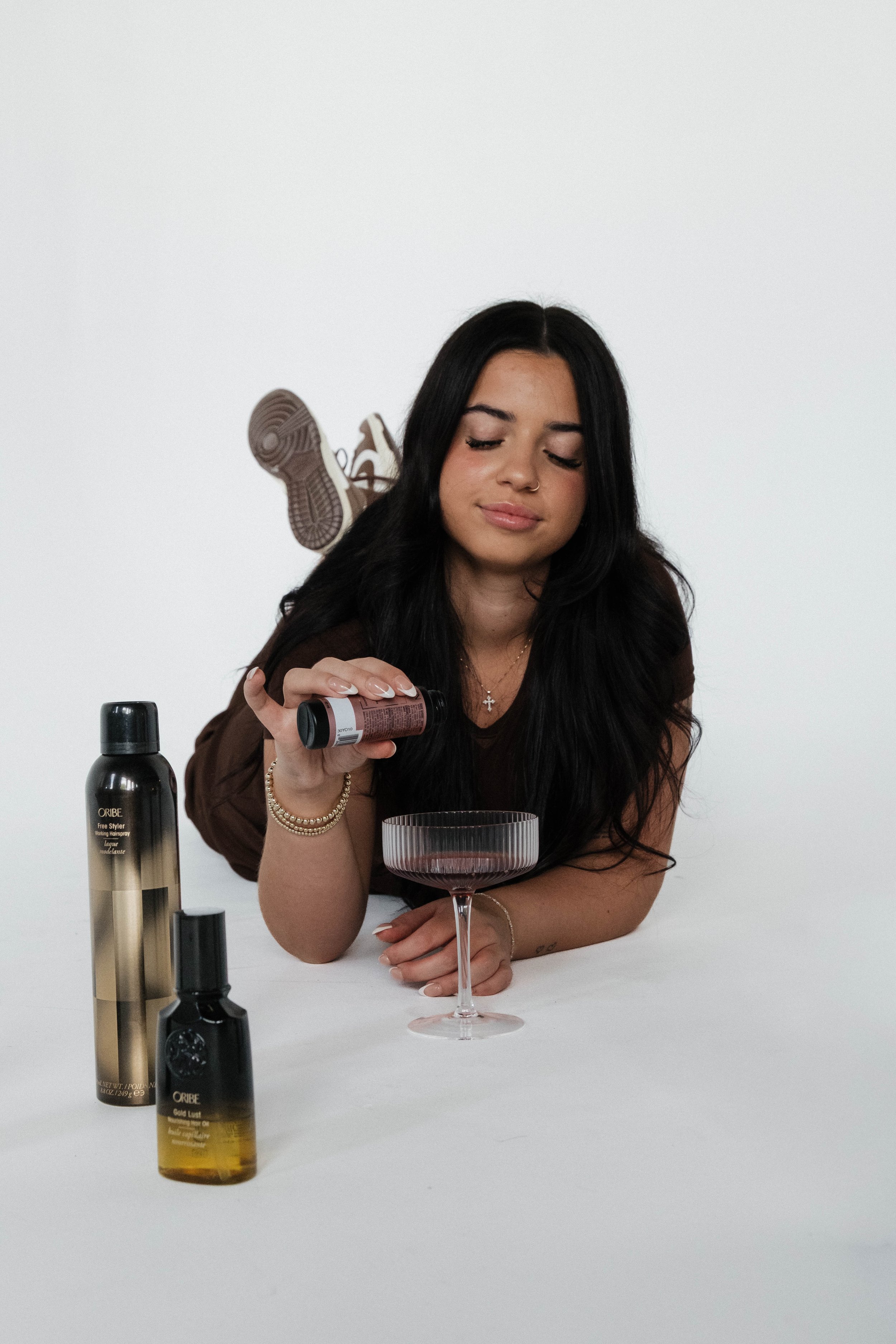 Syracuse, NY Branding photos, A woman with long black hair and a nose ring is lying on her stomach, pouring a bottle of liquor into a cocktail glass on a white table.