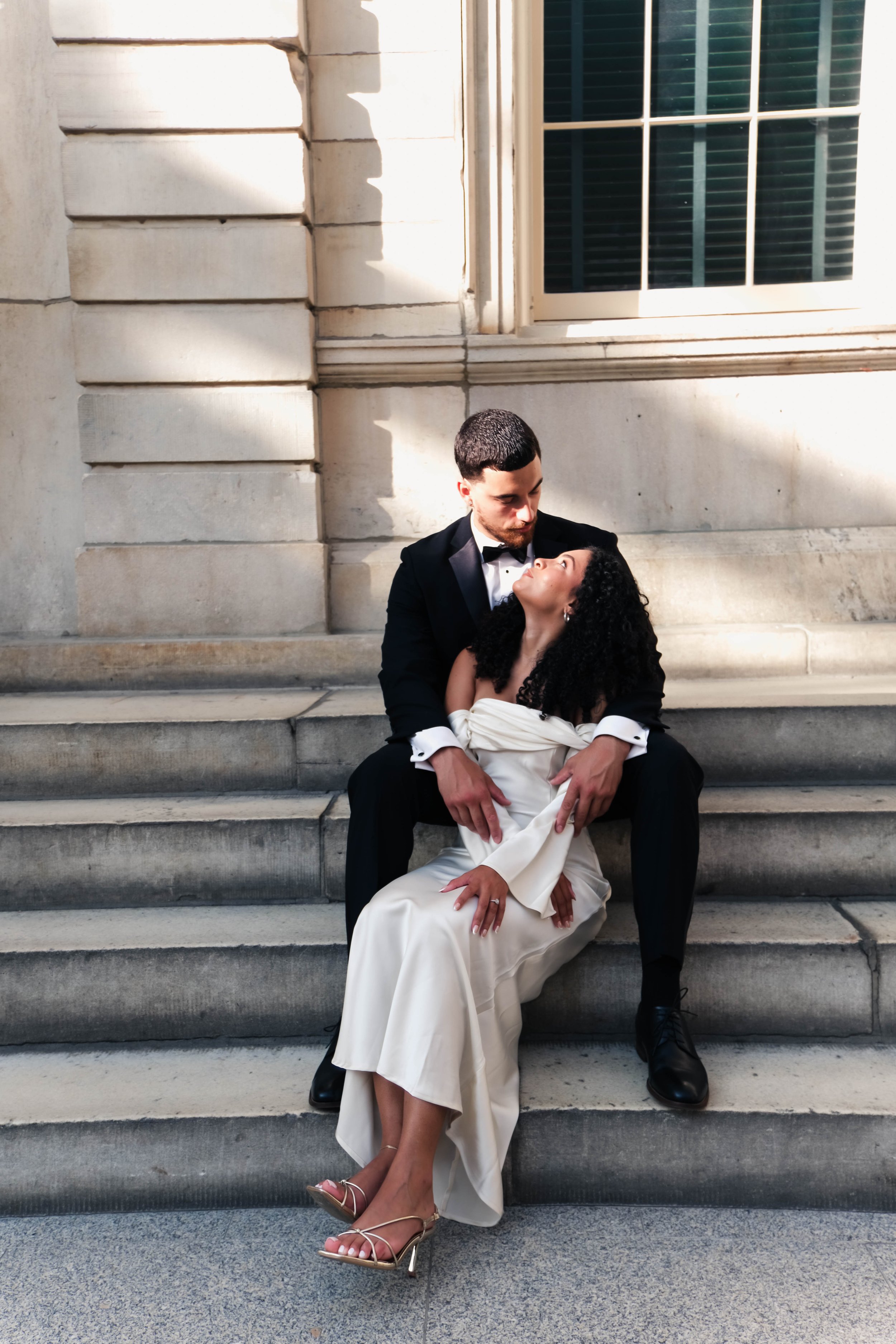 The Met Engagement Shoot  A newlywed couple sitting on stone steps outside a building, sharing an intimate moment, with the groom in a black tuxedo and the bride in a white wedding dress.