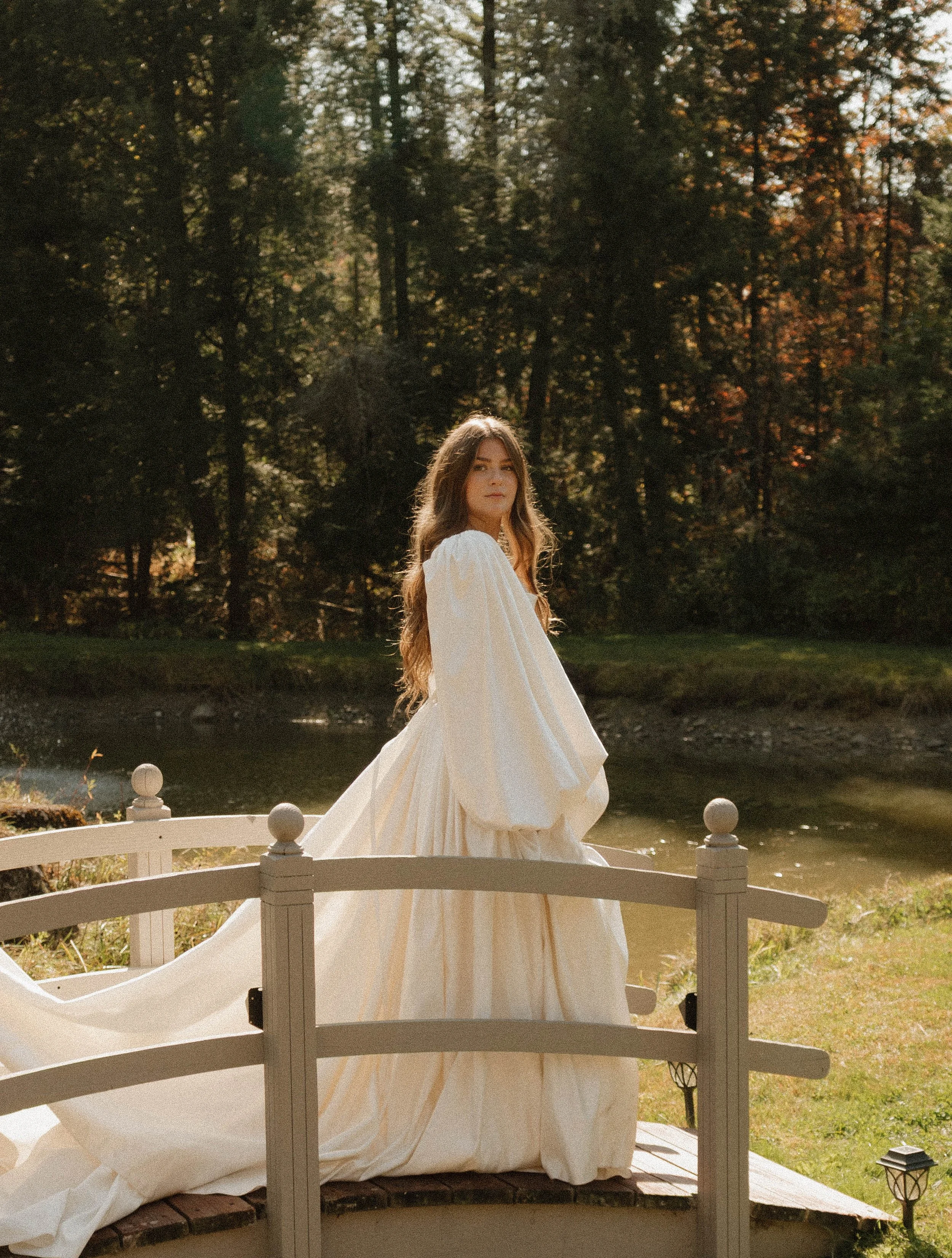 A woman in a flowing white dress standing on a small bridge near a body of water with trees in the background.