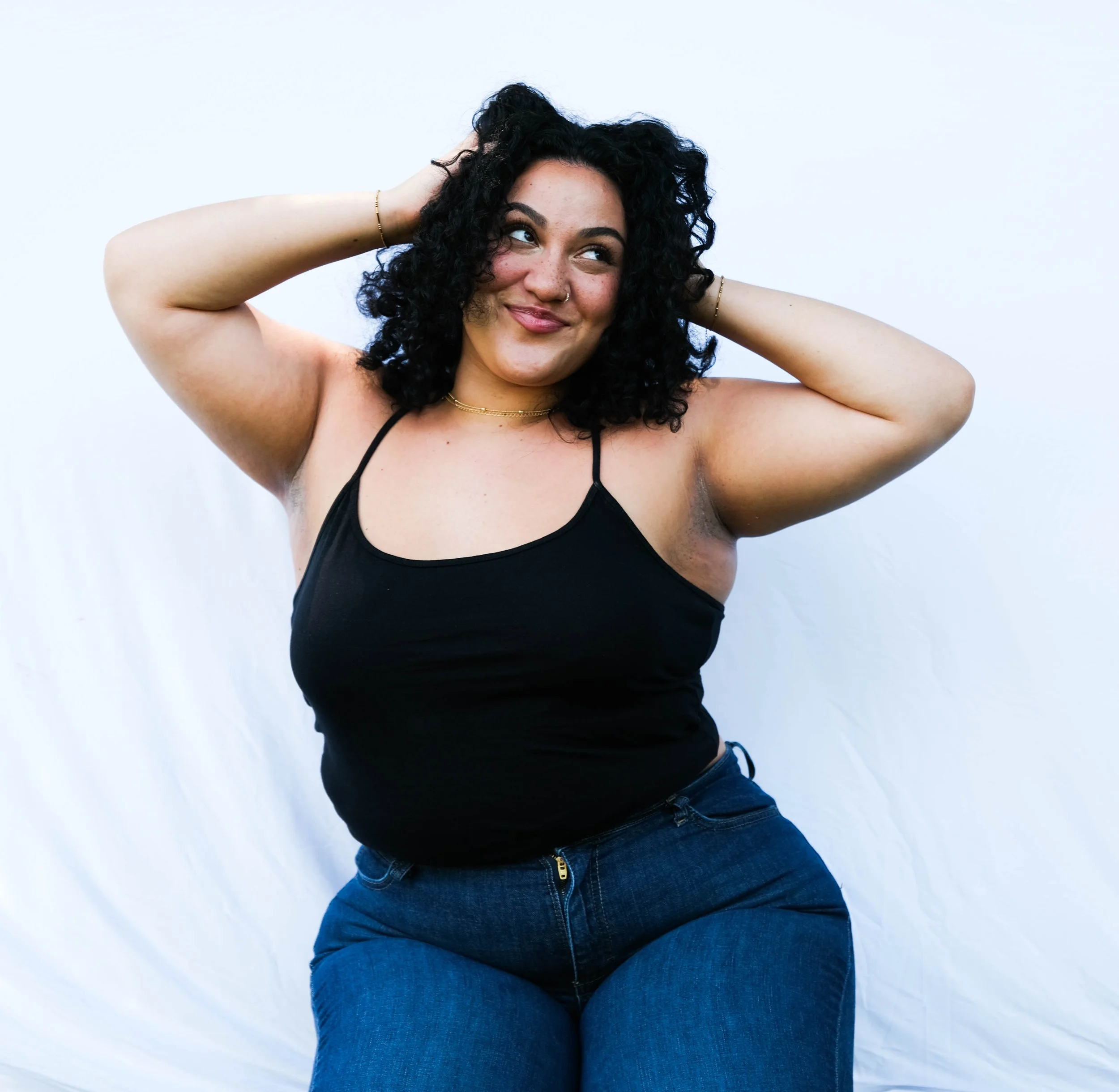 A woman with curly black hair wearing a black tank top and blue jeans, smiling and adjusting her hair against a white background.
