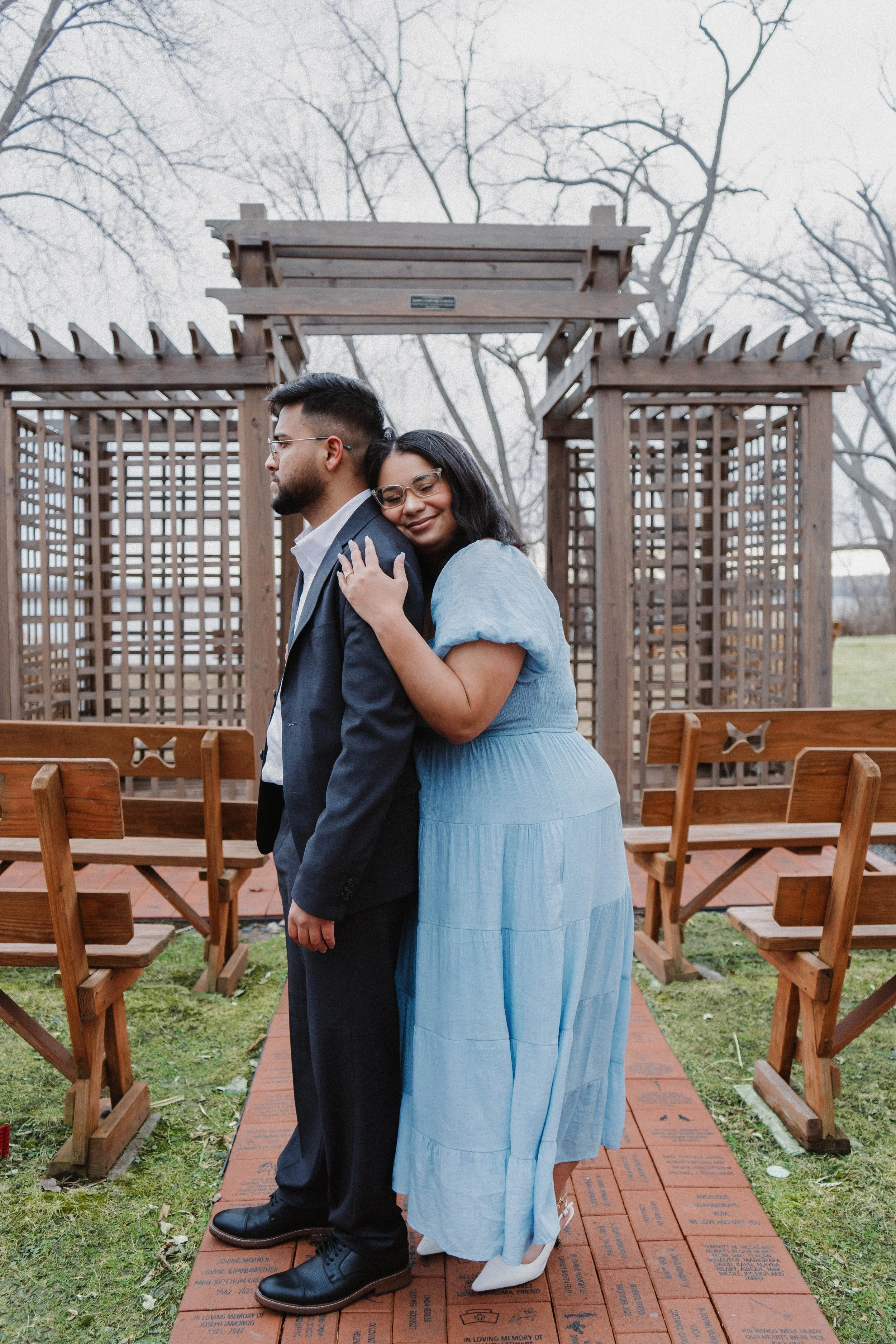 A woman in a blue dress hugging a man in a suit outdoors near a wooden structure with benches and leafless trees in the background.