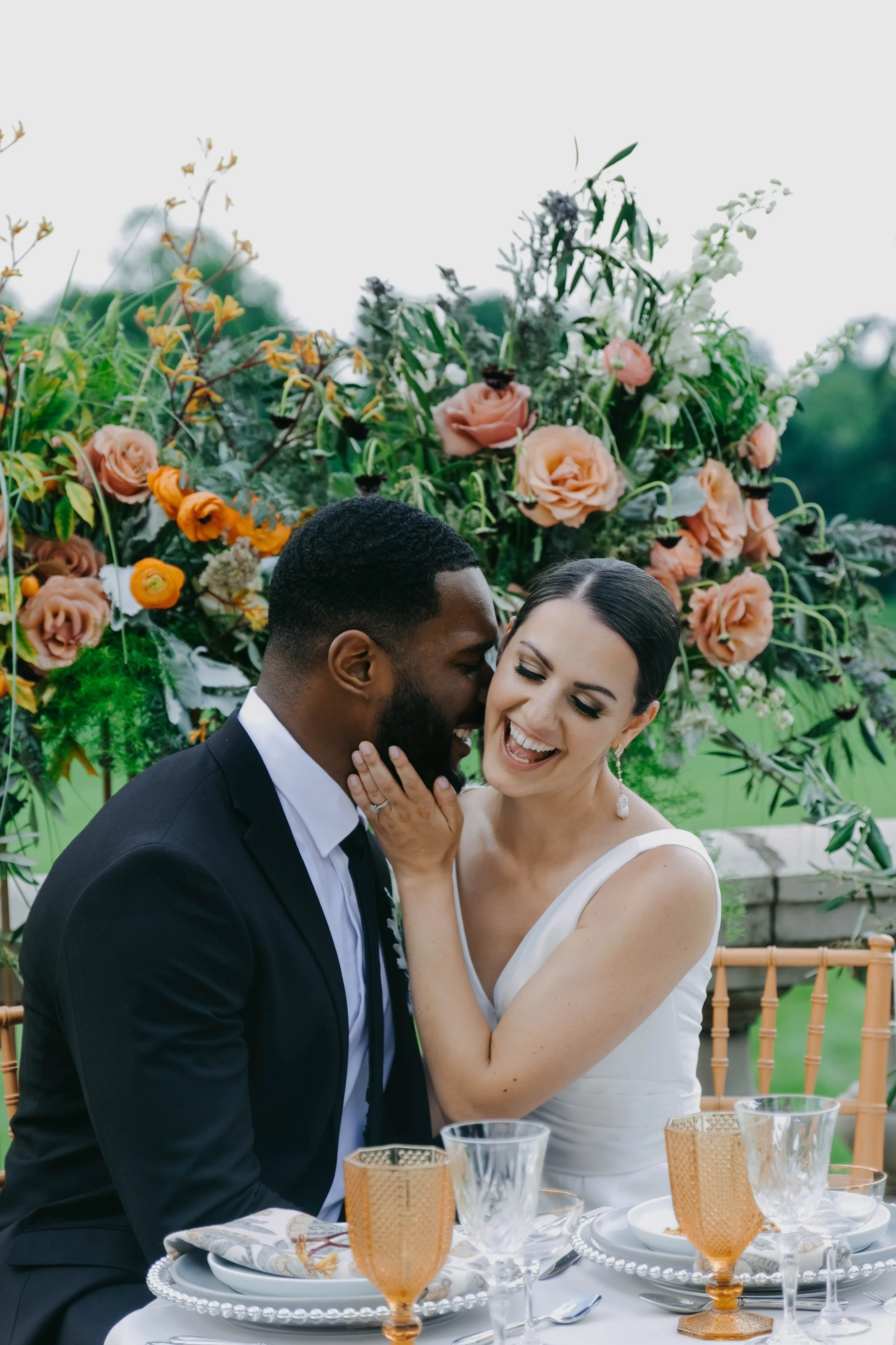 A smiling woman in a white dress touching her face to a man in a black suit, both enjoying an outdoor wedding during daytime with a large flower arrangement in the background.
