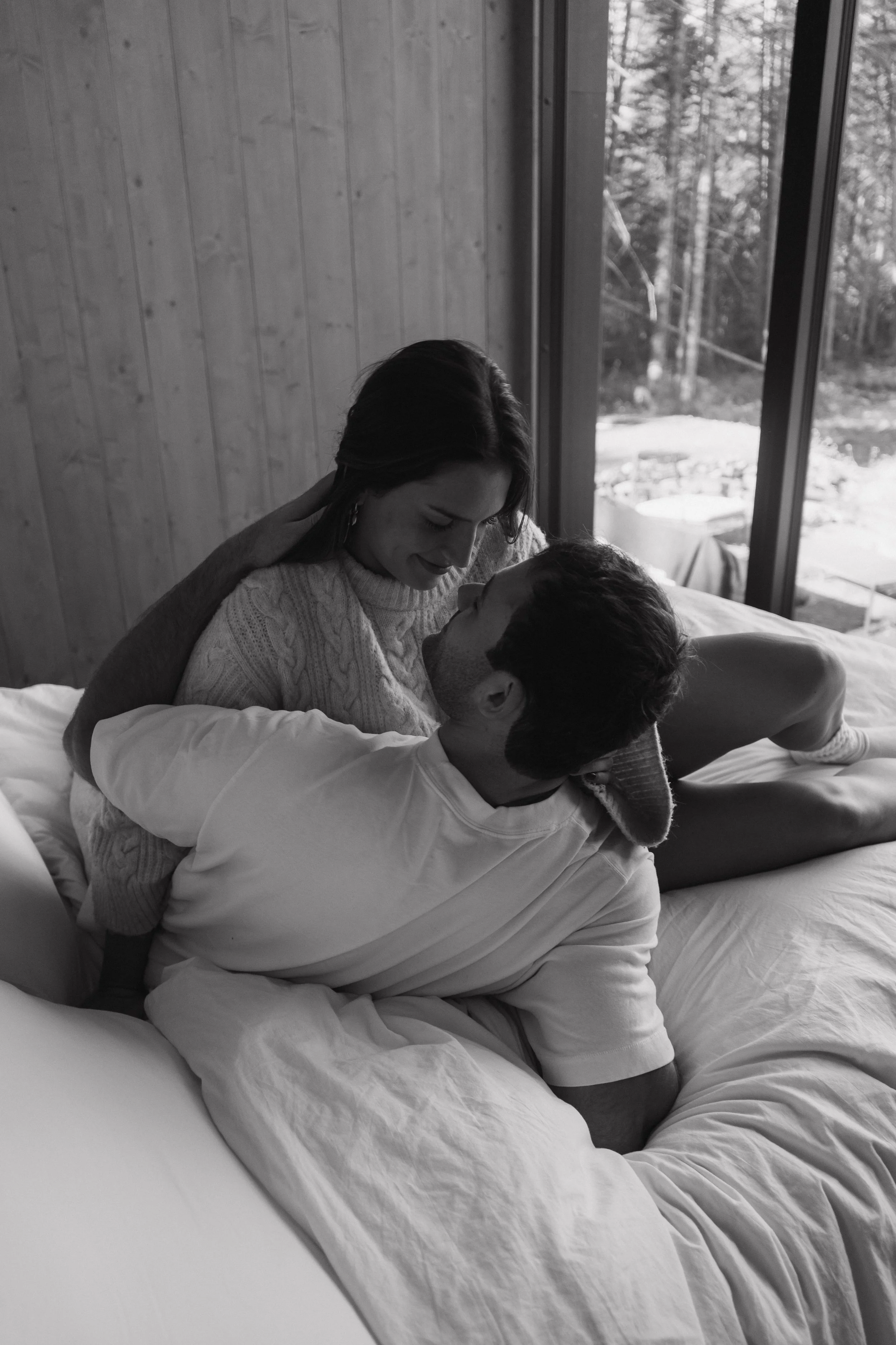 A black and white photo of a couple on a bed, with the woman on top and the man lying down, sharing an intimate moment inside a room with wooden walls and large windows showing an outdoor wooded landscape.