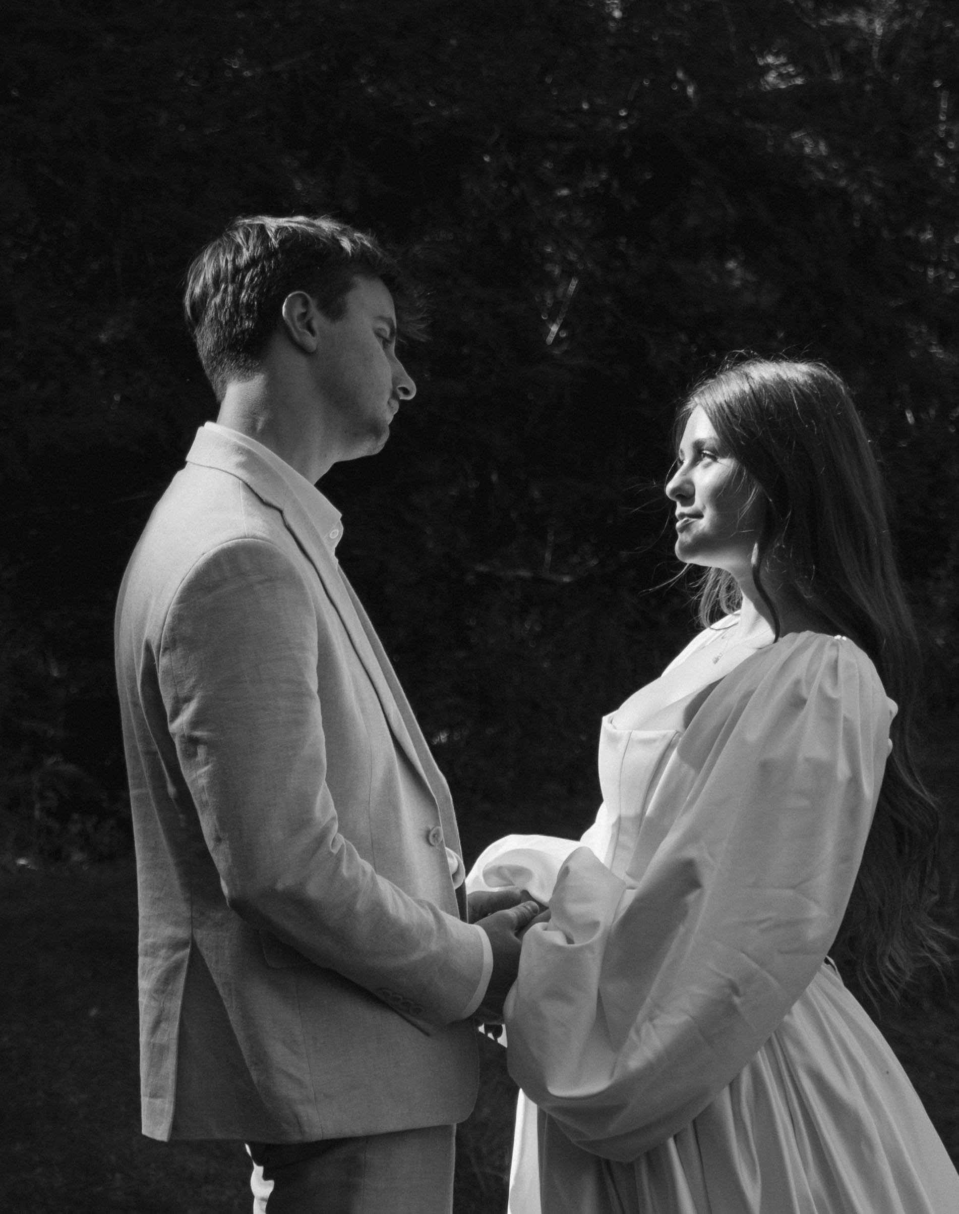 A black-and-white photo of a man and woman holding hands, standing face to face outdoors, looking at each other with a dark wooded background.